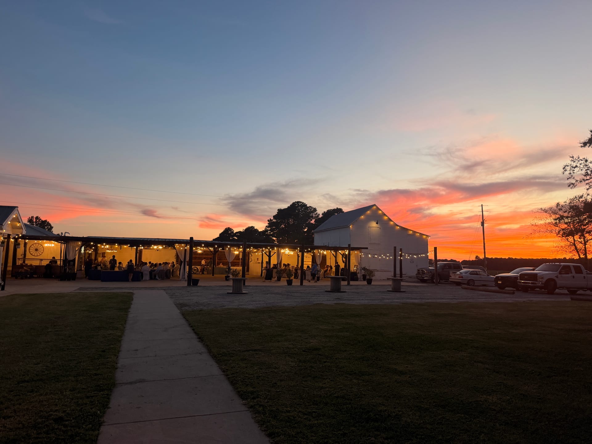 Guests gather under a warmly lit open-air pavilion at Scarborough House as a vivid orange and pink sunset fills the sky behind the white barn venue.