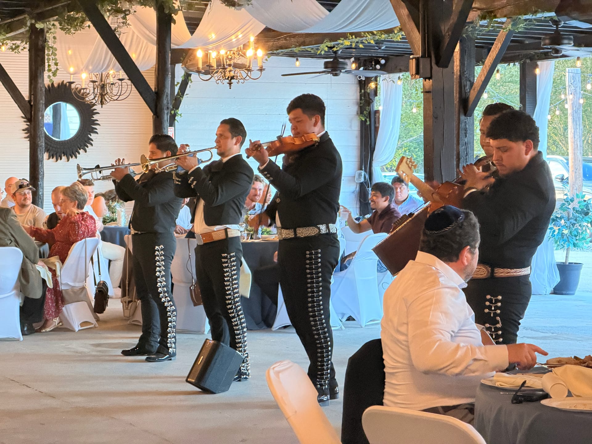 A mariachi band in traditional charro suits plays trumpet and violin for seated wedding guests inside a warmly lit decorated pavilion at Scarborough House.