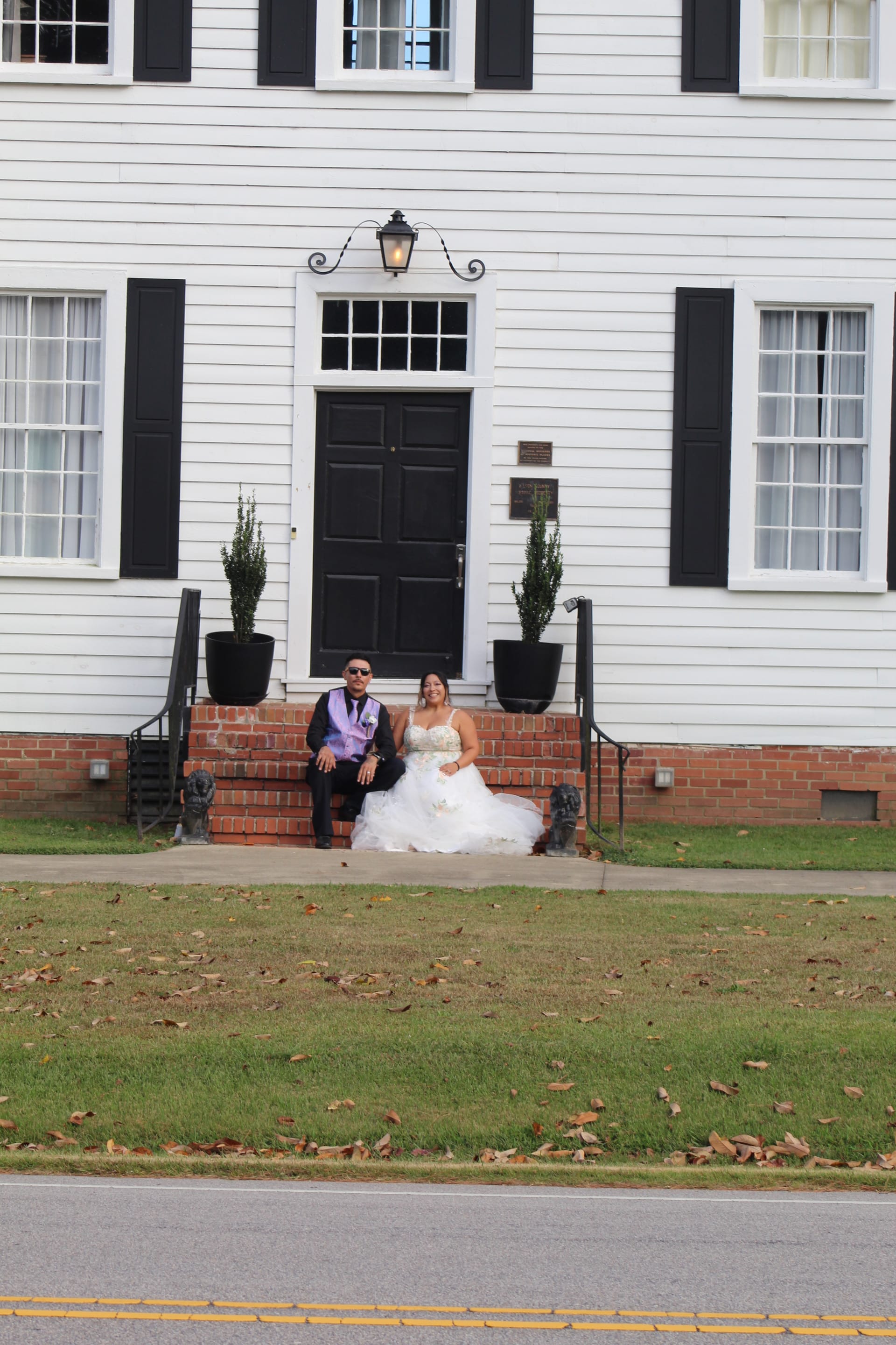 Bride in a white ball gown and groom in a dark suit with lavender vest sit together on the brick front steps of the 1821 Scarborough House historic manor.