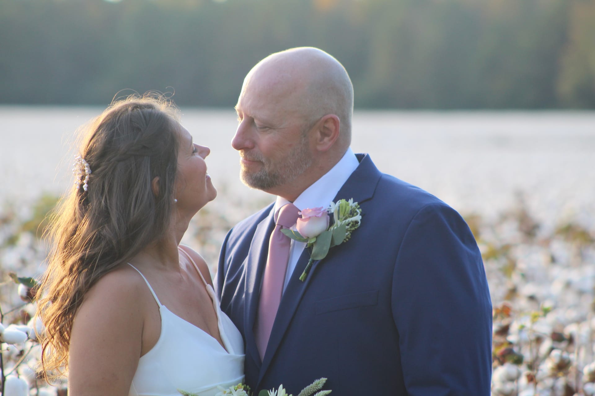 Bride in white gown and groom in navy suit with pink boutonniere share an intimate gaze in a sunlit cotton field at Scarborough House estate.