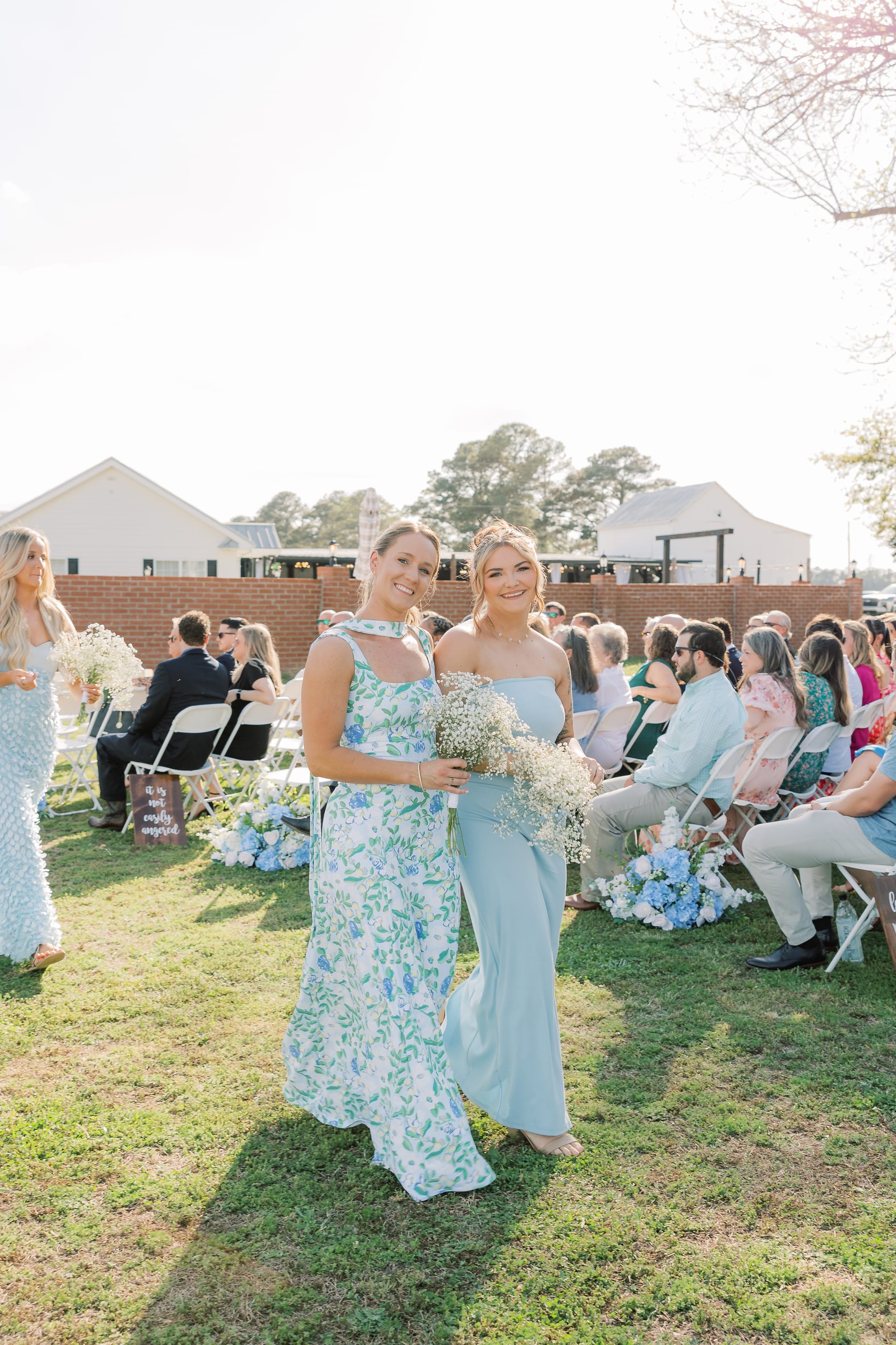 Two bridesmaids in a floral dress and light blue gown walk down the aisle carrying baby's breath bouquets during an outdoor wedding ceremony at Scarborough House.