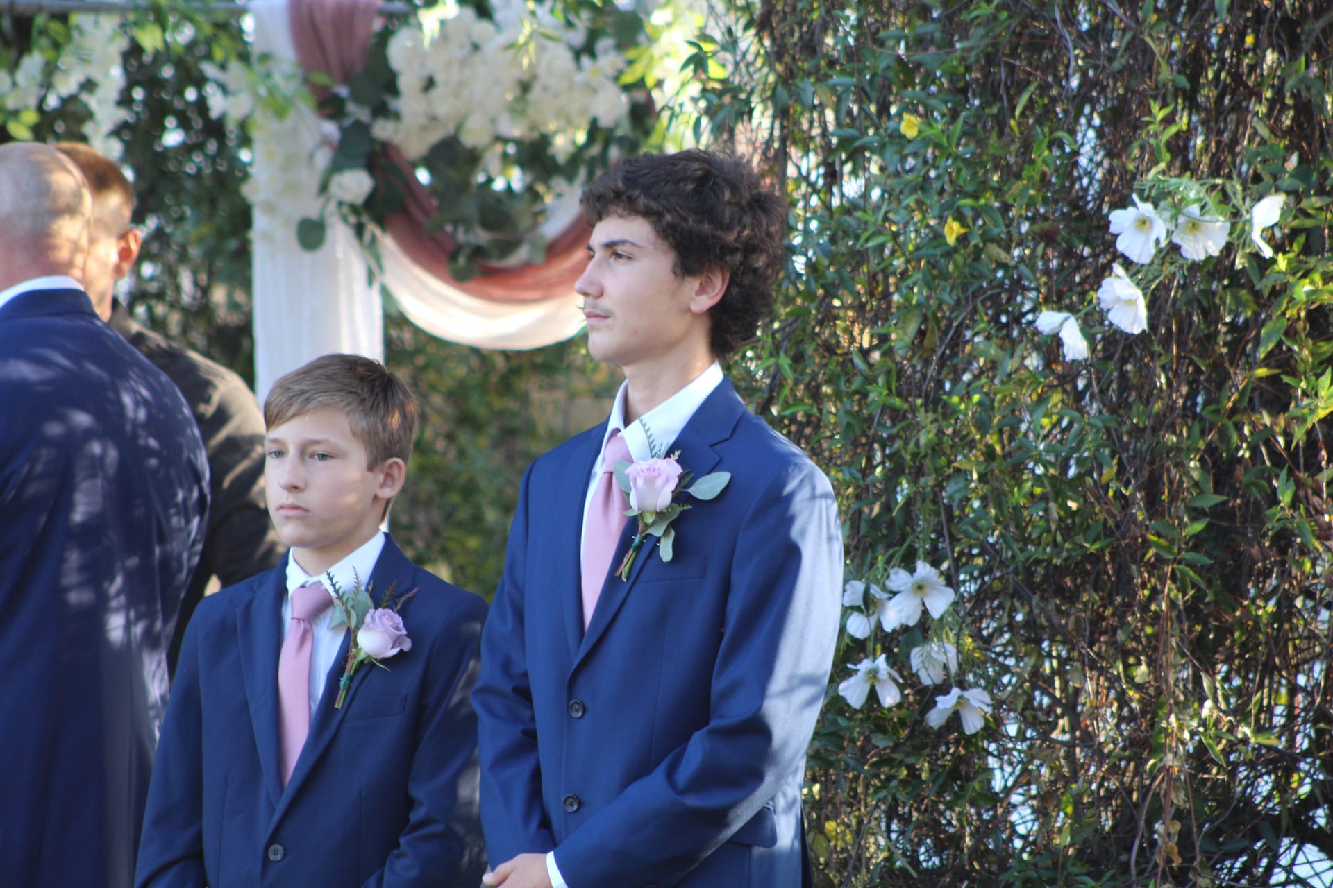 Two young groomsmen in navy suits and pink ties with rose boutonnieres stand attentively during an outdoor wedding ceremony at Scarborough House, with a floral arch and white blooms visible in the background.
