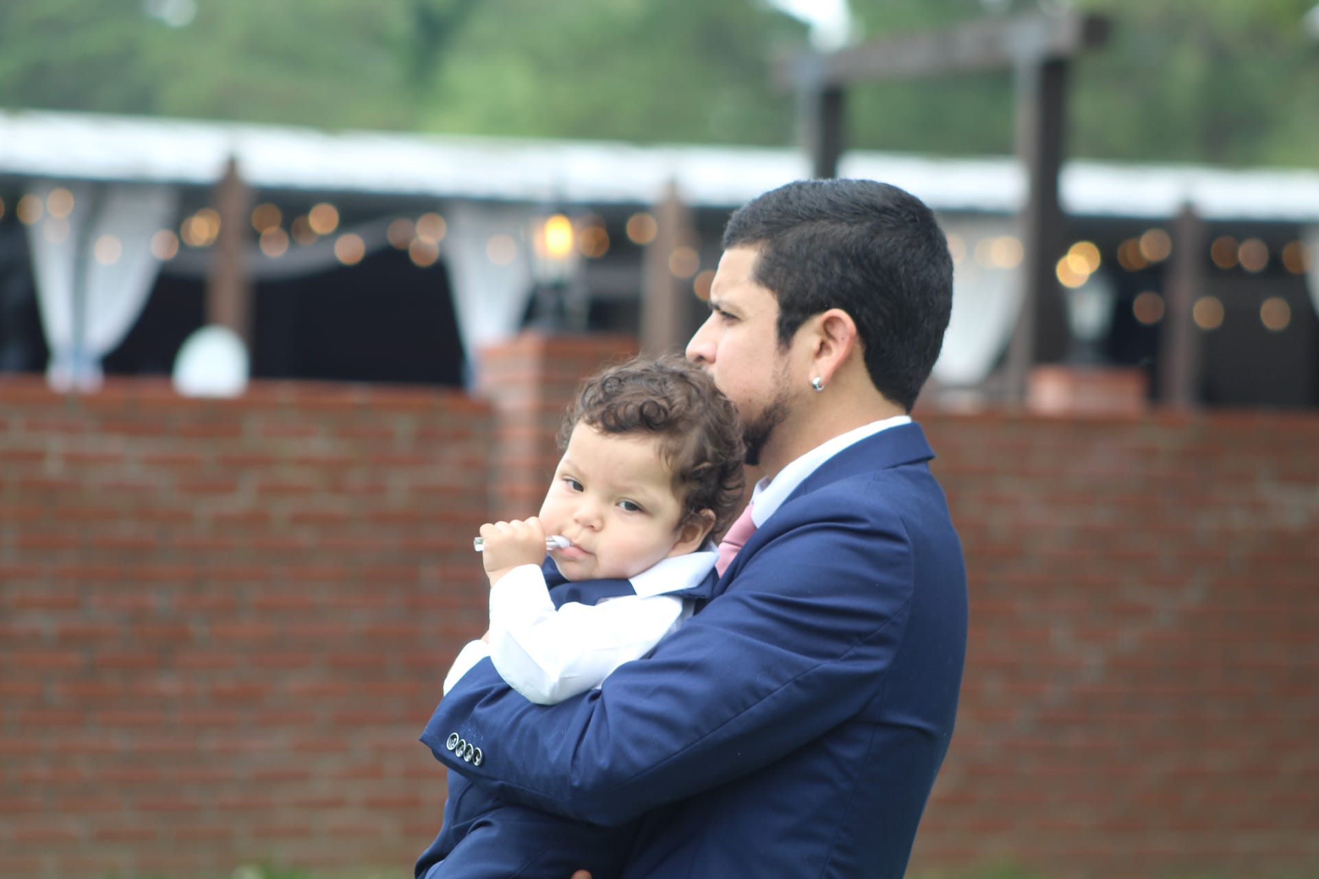 A man in a navy suit holds a young toddler dressed in formal attire at an outdoor wedding venue with a brick wall and festoon-lit tent in the background.