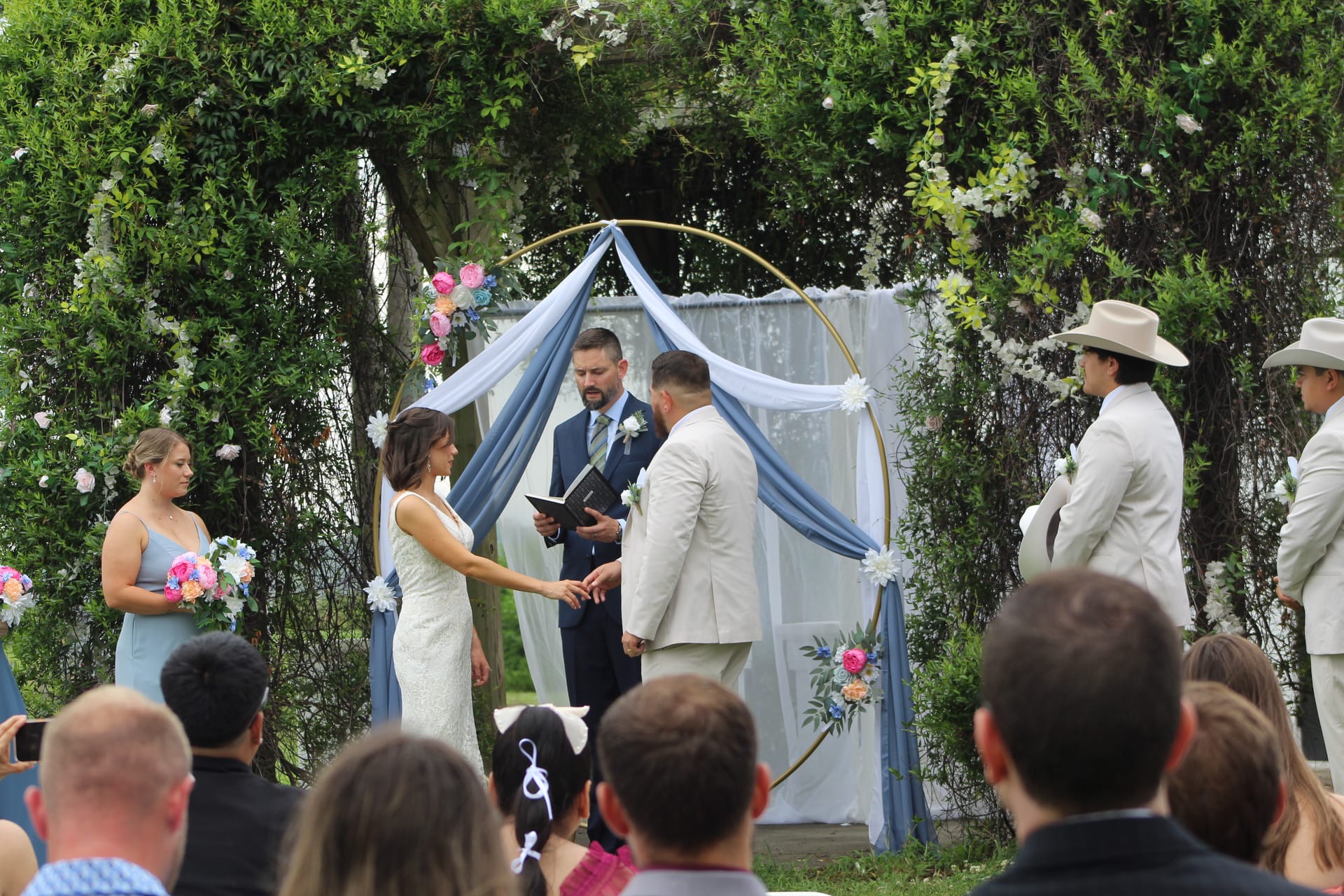 Bride and groom exchange rings beneath a gold circular arch draped in dusty blue fabric and colorful florals surrounded by lush greenery at an outdoor estate wedding ceremony.