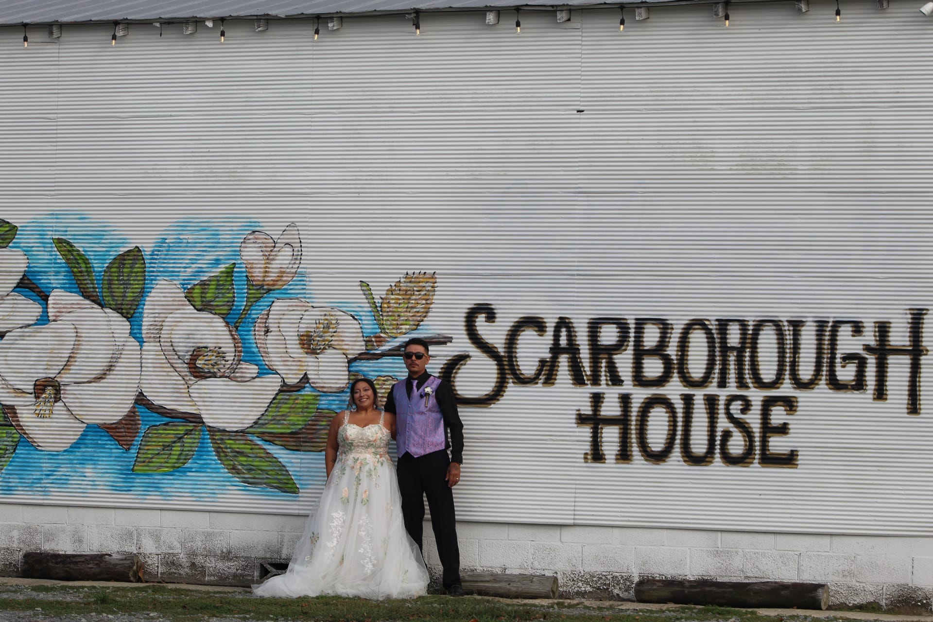 Bride in a floral ball gown and groom in a purple vest and black suit pose together in front of the painted magnolia mural and Scarborough House sign on the exterior barn wall.