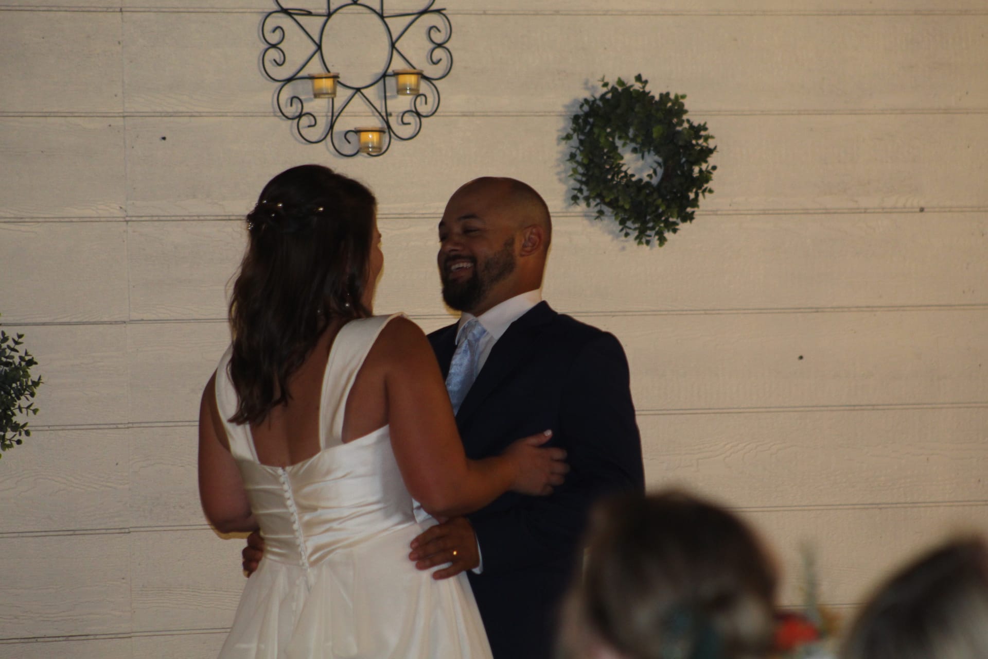 Smiling groom in a navy suit shares a first dance with his bride in a white gown inside a warmly lit venue decorated with a wreath and candlelit wall sconce.