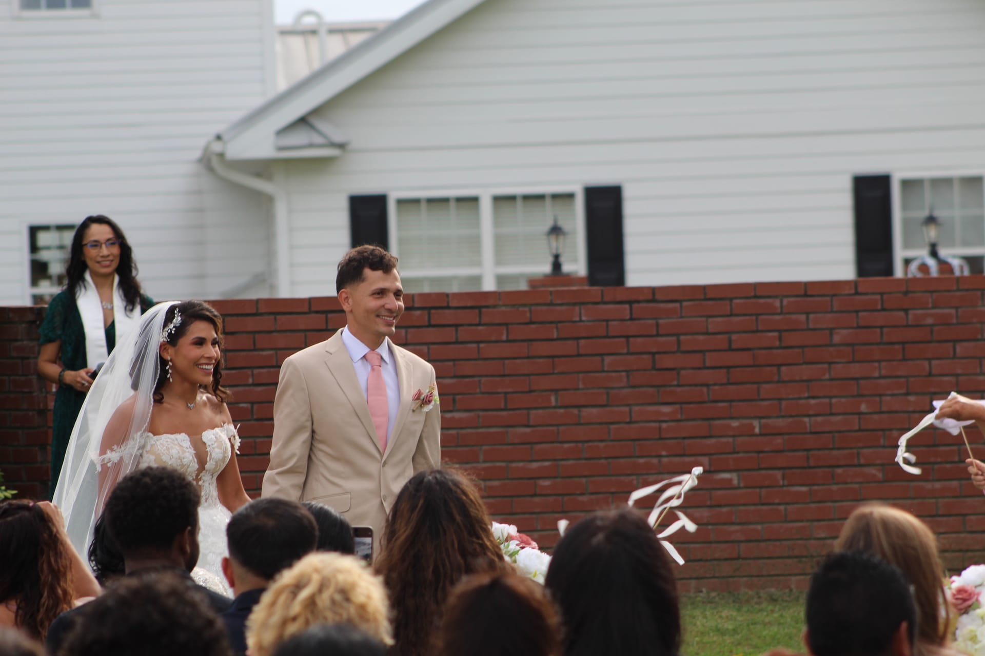 A smiling bride in a white lace gown and veil stands beside her groom in a tan suit with pink tie during an outdoor wedding ceremony at Scarborough House historic estate, with the 1821 white manor house visible in the background.