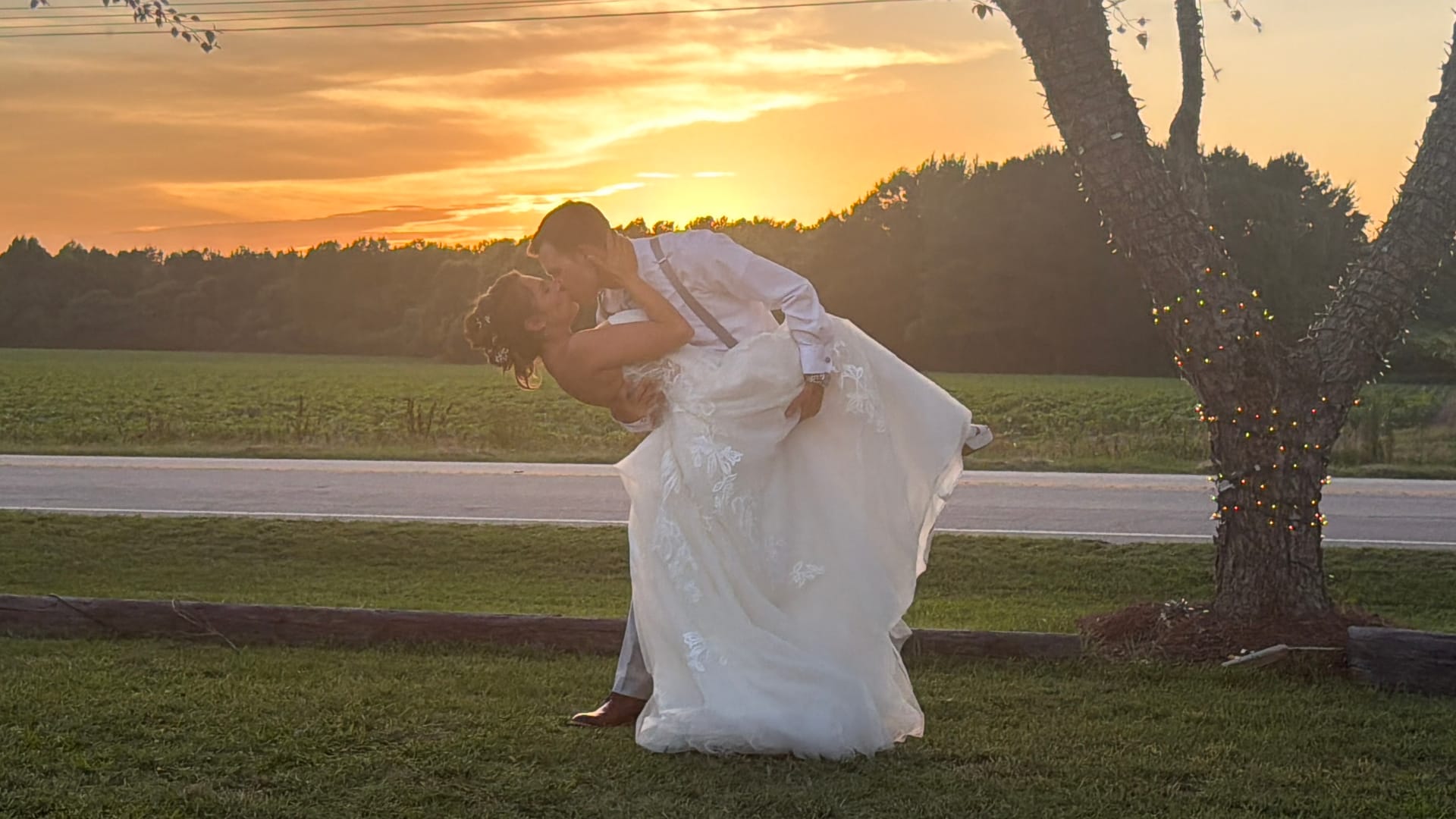 Bride and groom share a romantic dip kiss silhouetted against a vivid golden sunset beside a fairy-light-wrapped oak tree at Scarborough House estate wedding venue.