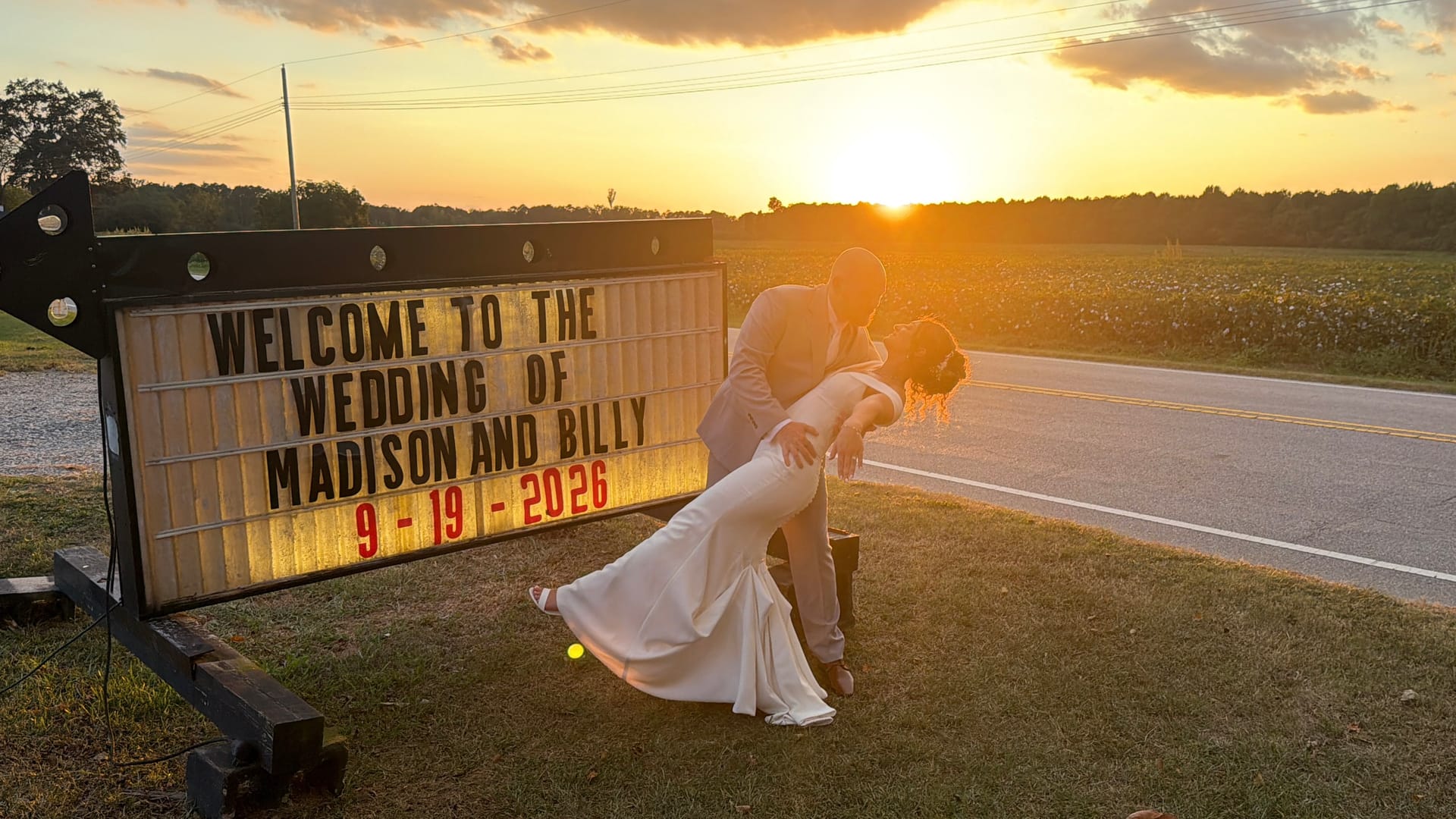 Bride and groom share a romantic dip kiss silhouetted against a vibrant golden sunset beside a lit marquee sign welcoming guests to the wedding of Madison and Billy on 9-19-2026.