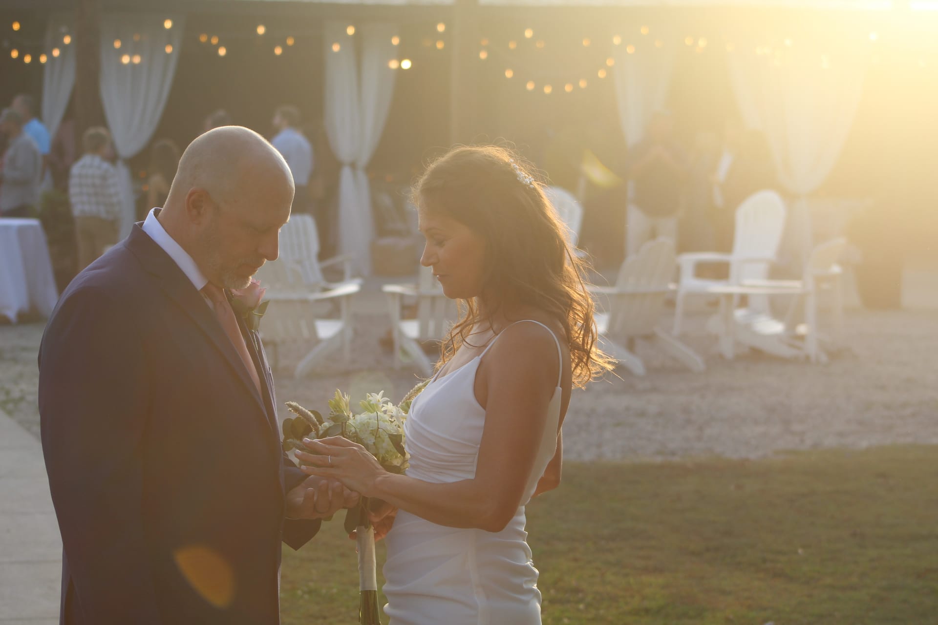 Bride and groom exchange rings during an outdoor golden-hour wedding ceremony at Scarborough House, bathed in warm sunset light with string lights and white chairs softly blurred in the background.