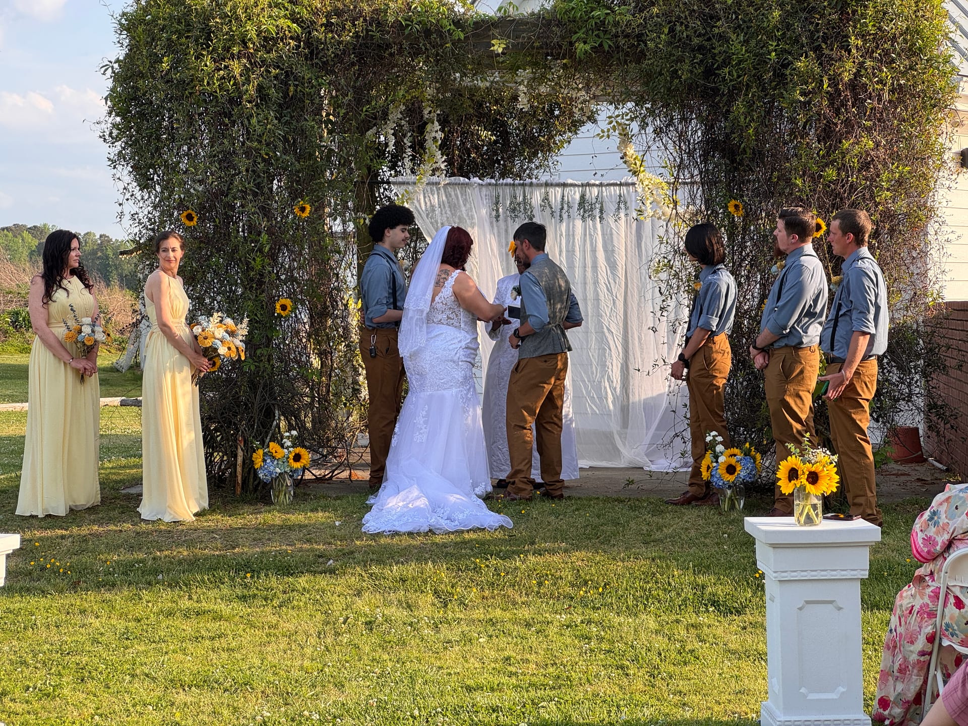 Bride and groom exchange vows beneath a lush greenery arch decorated with sunflowers and white drapery on the ceremony lawn at Scarborough House, flanked by bridesmaids in yellow gowns and groomsmen in denim and tan.