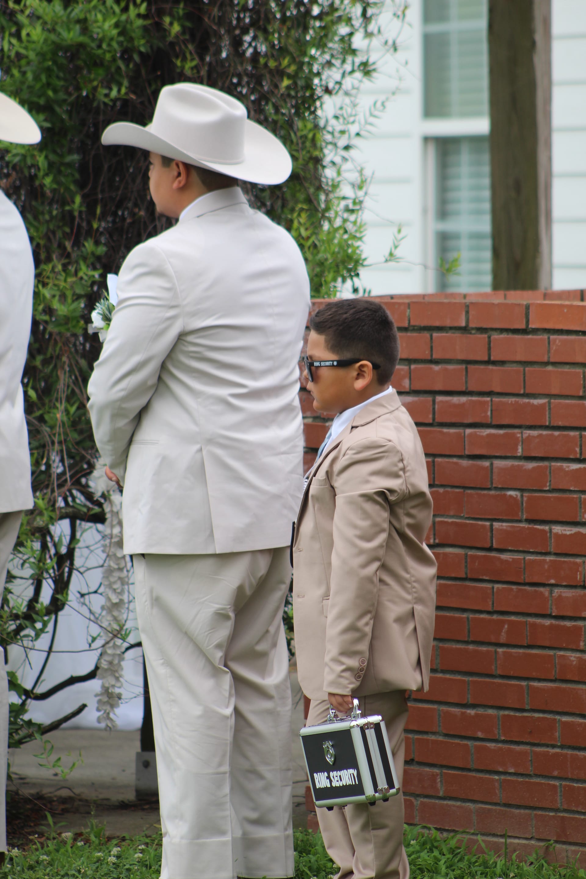 A young ring bearer wearing sunglasses and a tan suit holds a Ring Security briefcase while standing beside a groomsman in a white suit and cowboy hat at an outdoor wedding ceremony.