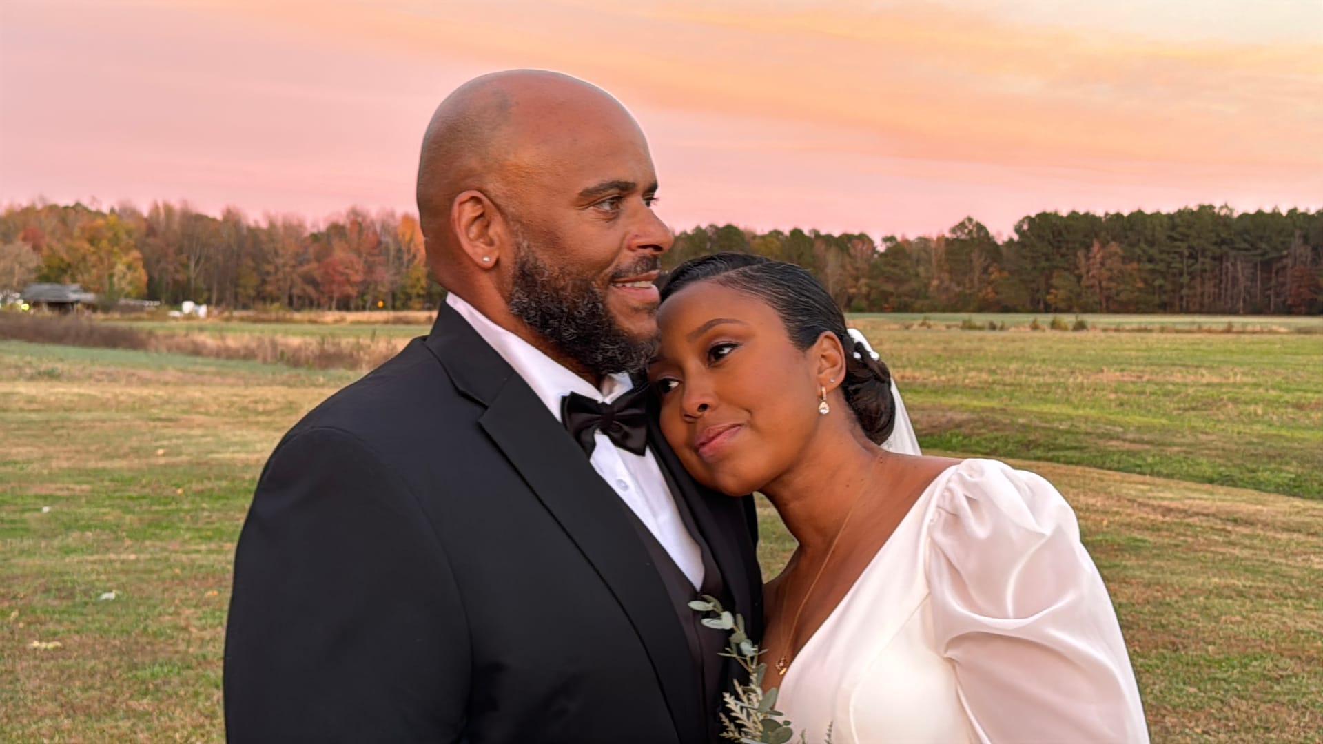 Bride in a white puff-sleeve gown rests her head on the groom's shoulder as they share a tender moment in an open farmland field beneath a glowing pink and orange sunset sky at Scarborough House.