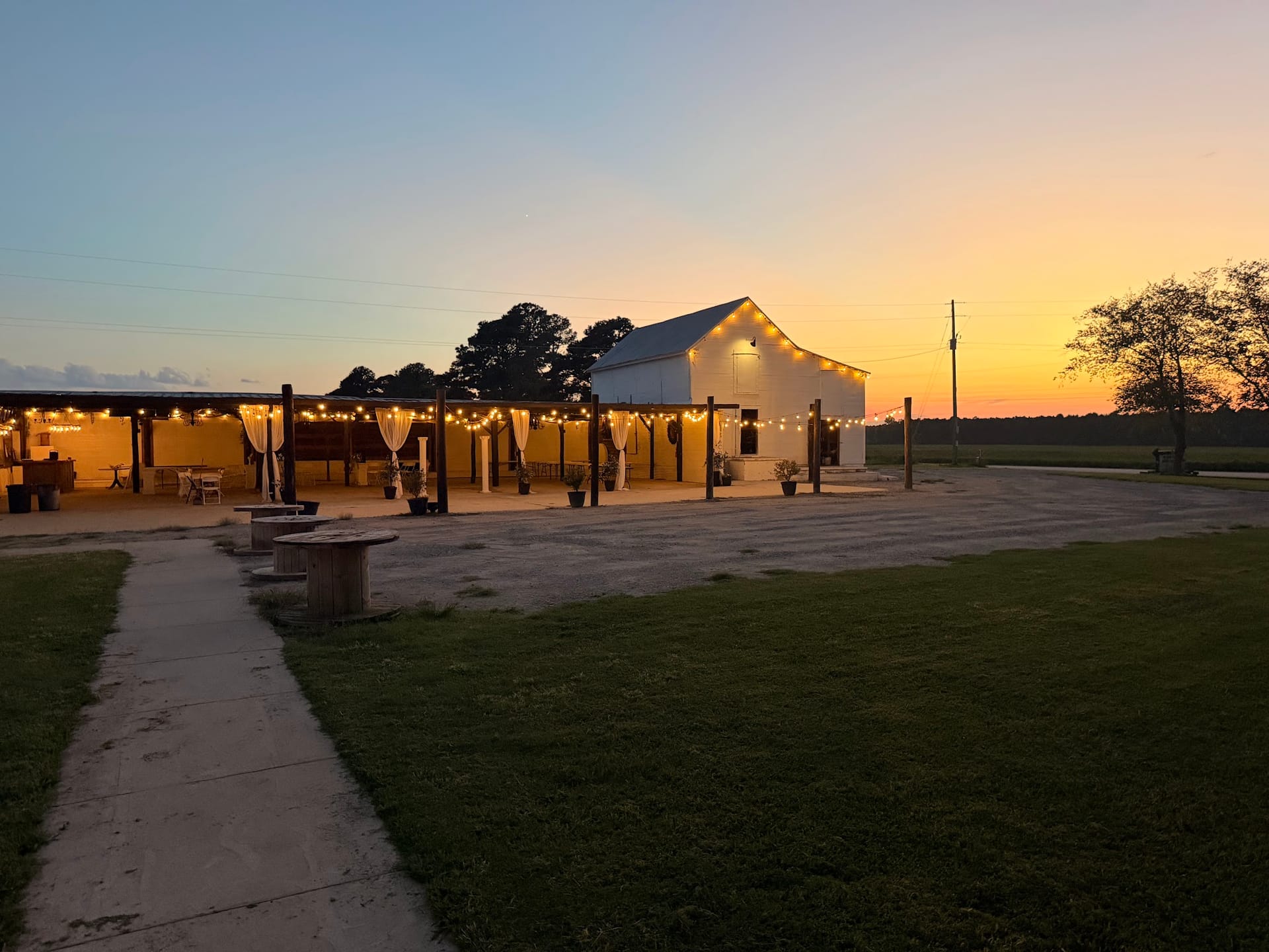 A white barn wedding venue glows warmly under string lights at sunset with a covered outdoor pavilion and open farmland fields in the background.