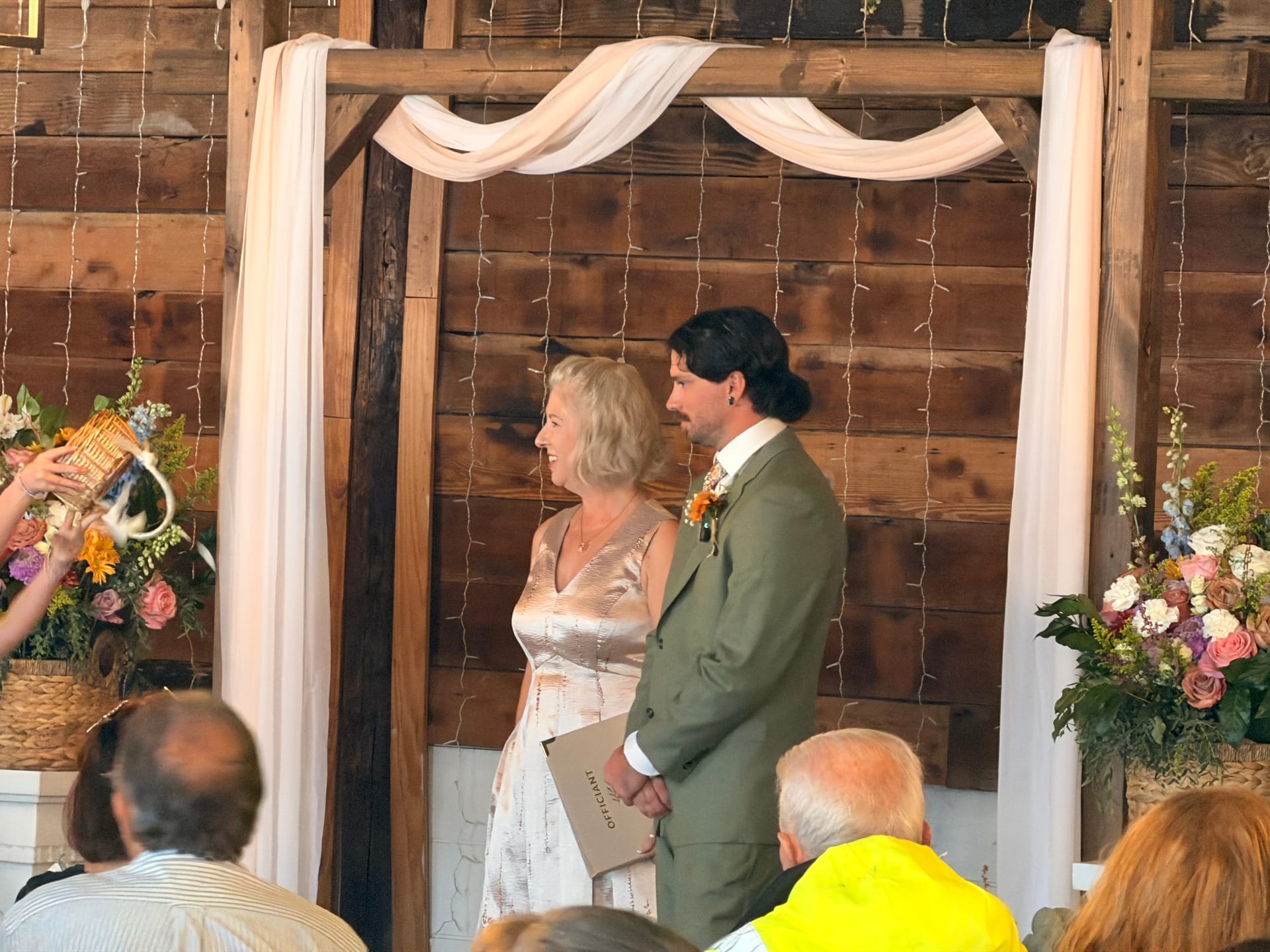 Bride in a champagne dress and groom in an olive suit share a tender glance beneath a draped wooden arch adorned with fairy lights and wildflower arrangements during their barn wedding ceremony.