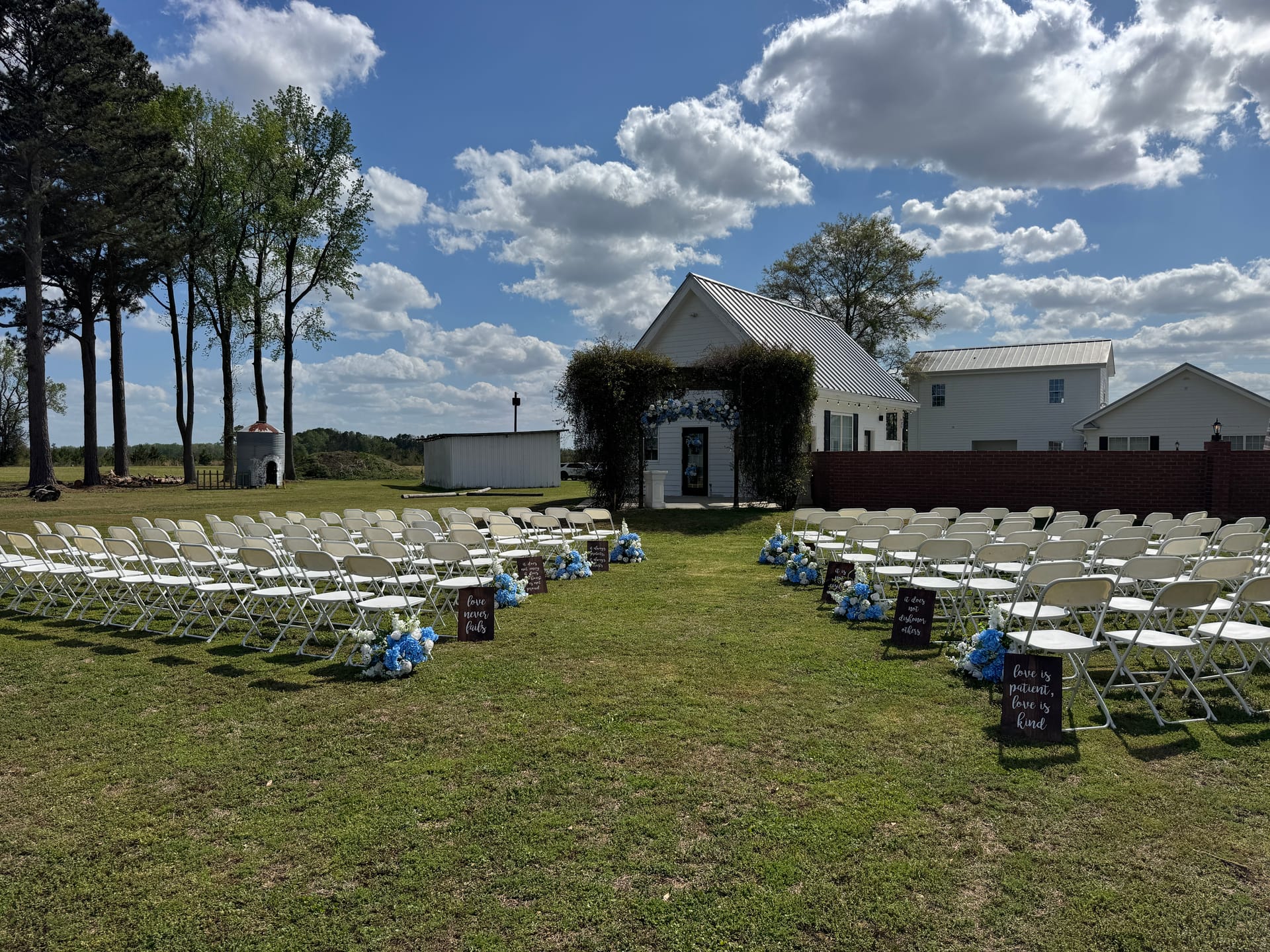 Outdoor wedding ceremony setup at Scarborough House featuring rows of white folding chairs flanking a floral-lined aisle with blue hydrangea arrangements and calligraphy signs leading to an ivy-draped white chapel entrance under a bright blue sky.