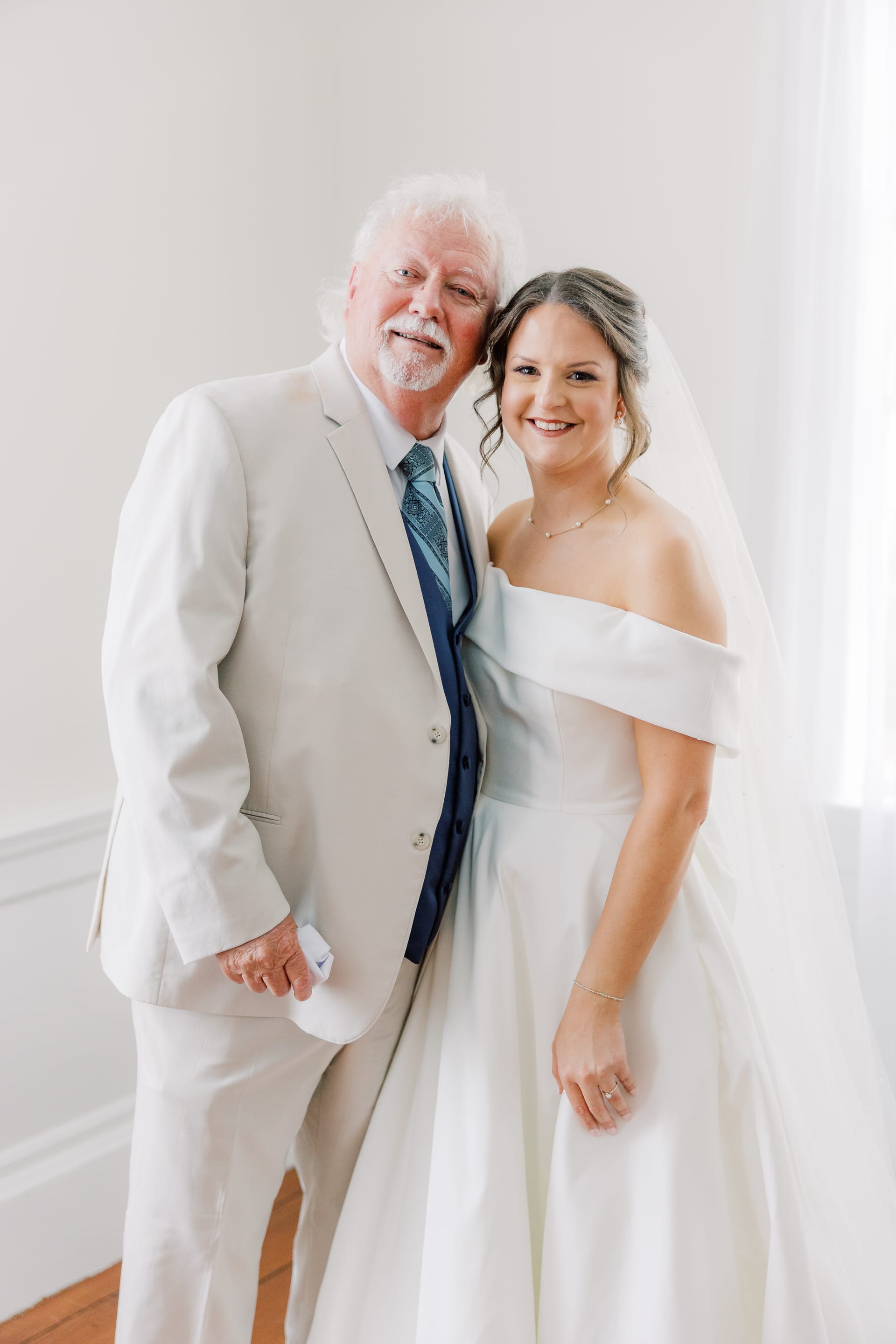 A smiling bride in an off-the-shoulder white gown and veil poses cheek-to-cheek with an older gentleman in a cream suit and blue tie inside Scarborough House.
