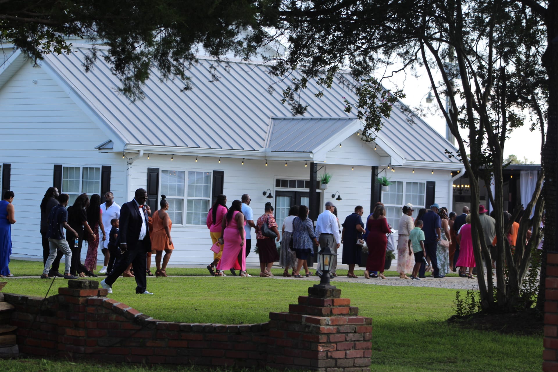 Wedding guests dressed in colorful formal attire walking toward the white farmhouse manor at Scarborough House as string lights glow at dusk.