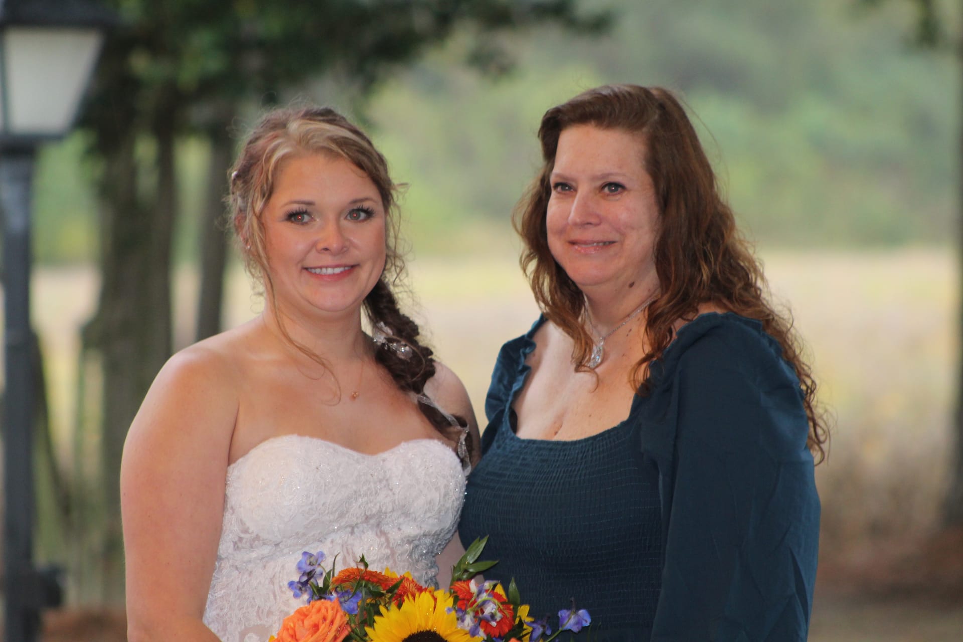 Bride in a strapless white lace gown holding a sunflower and wildflower bouquet poses with a guest in a teal dress against a soft bokeh backdrop of oak trees and farmland fields at Scarborough House.