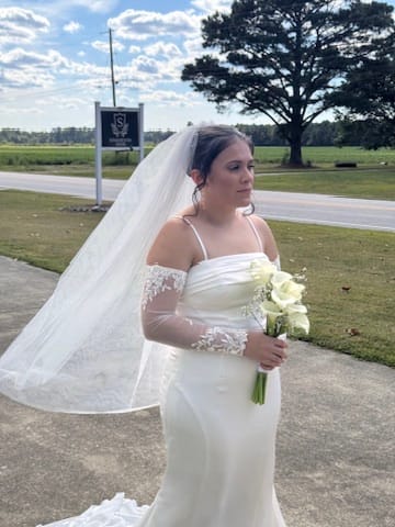 Bride in a fitted white lace-sleeve gown holding a white calla lily bouquet stands near the Scarborough House marquee sign with green lawns and farmland fields in the background.
