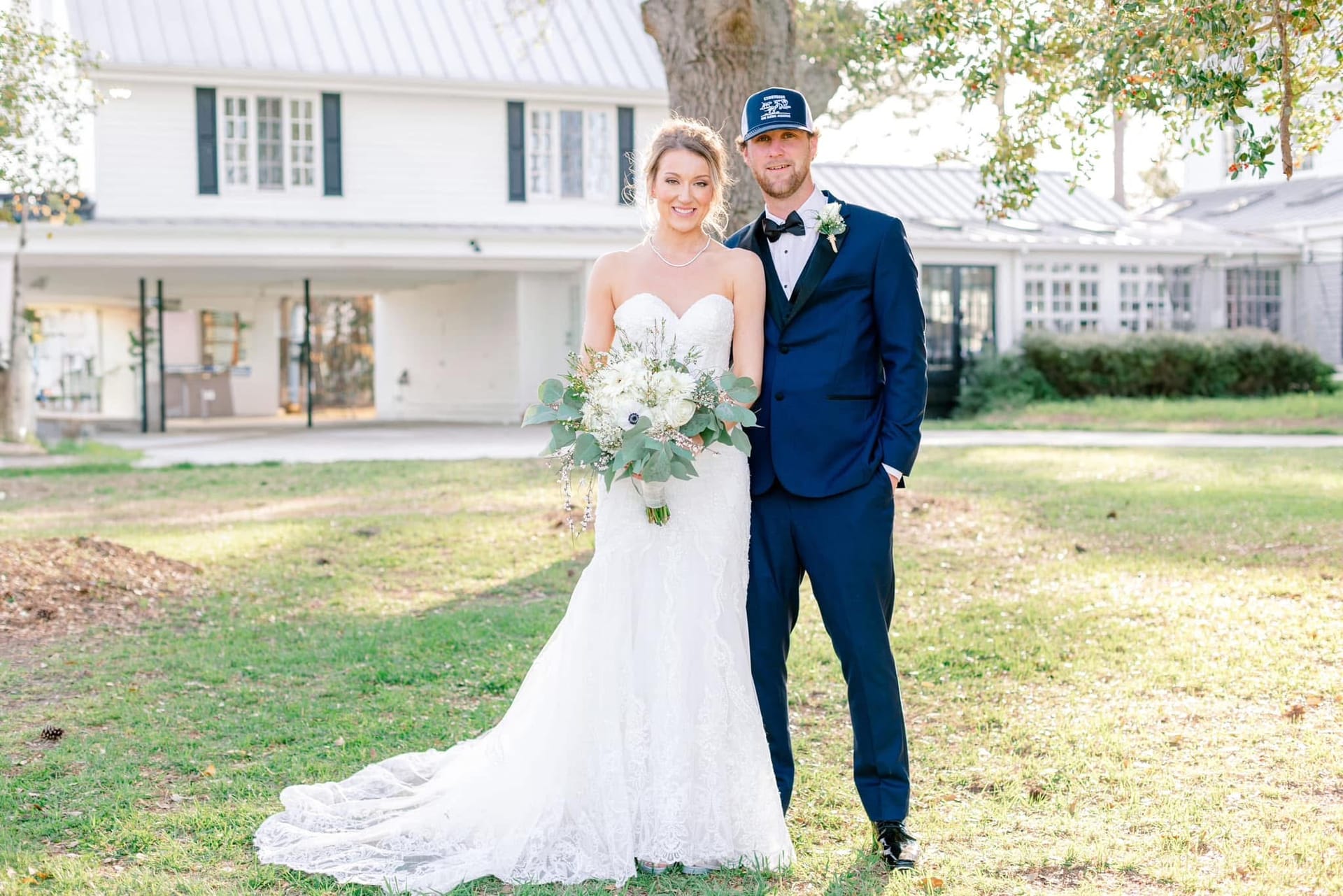 Bride in a lace strapless gown holding a white and greenery bouquet stands beside a suited groom wearing a navy tuxedo and trucker cap in front of the historic 1821 Scarborough House manor on a sunny day.