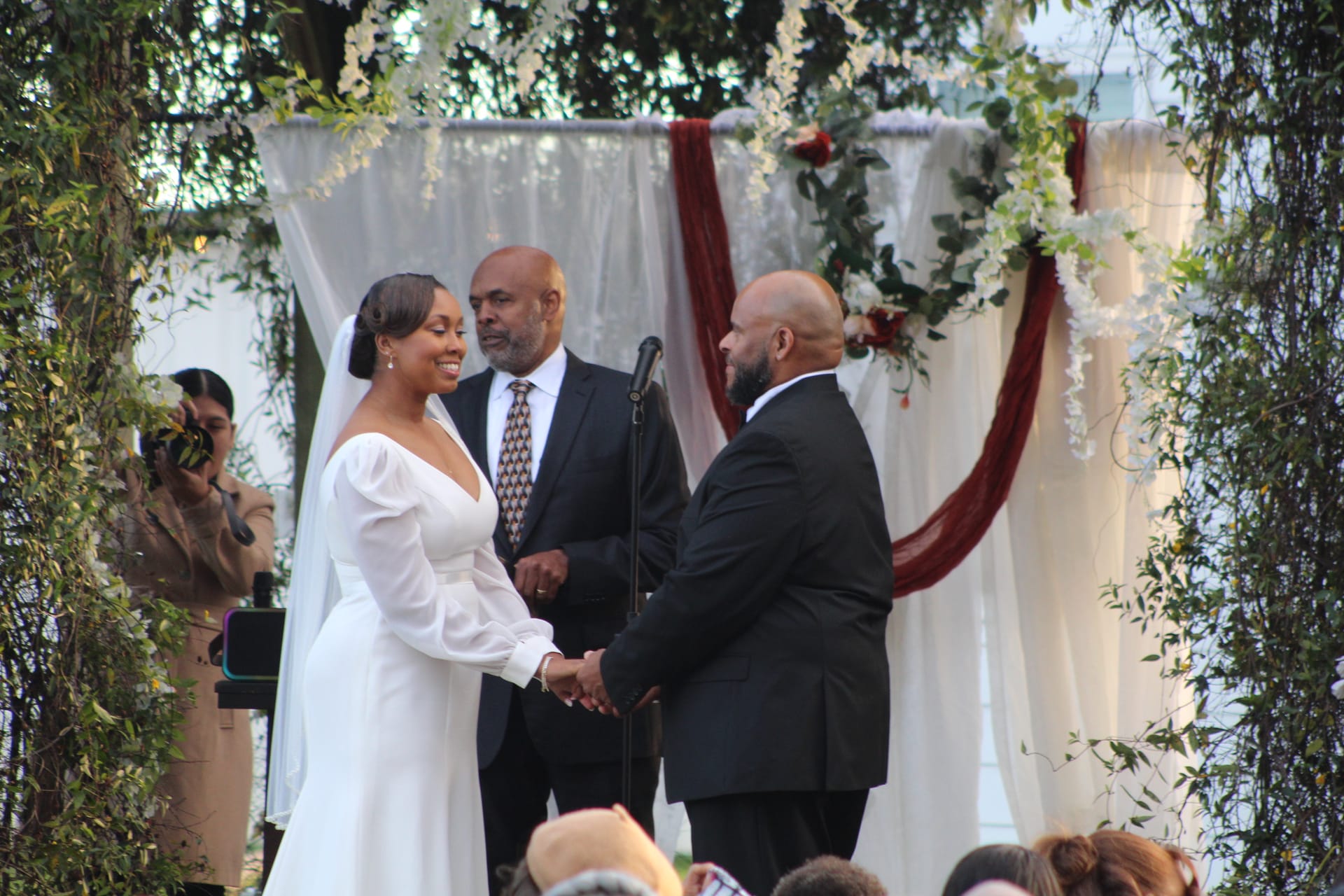 Bride in a long-sleeve white gown and groom in a black suit hold hands and exchange vows before an officiant beneath a floral and draped altar on the Scarborough House ceremony lawn.