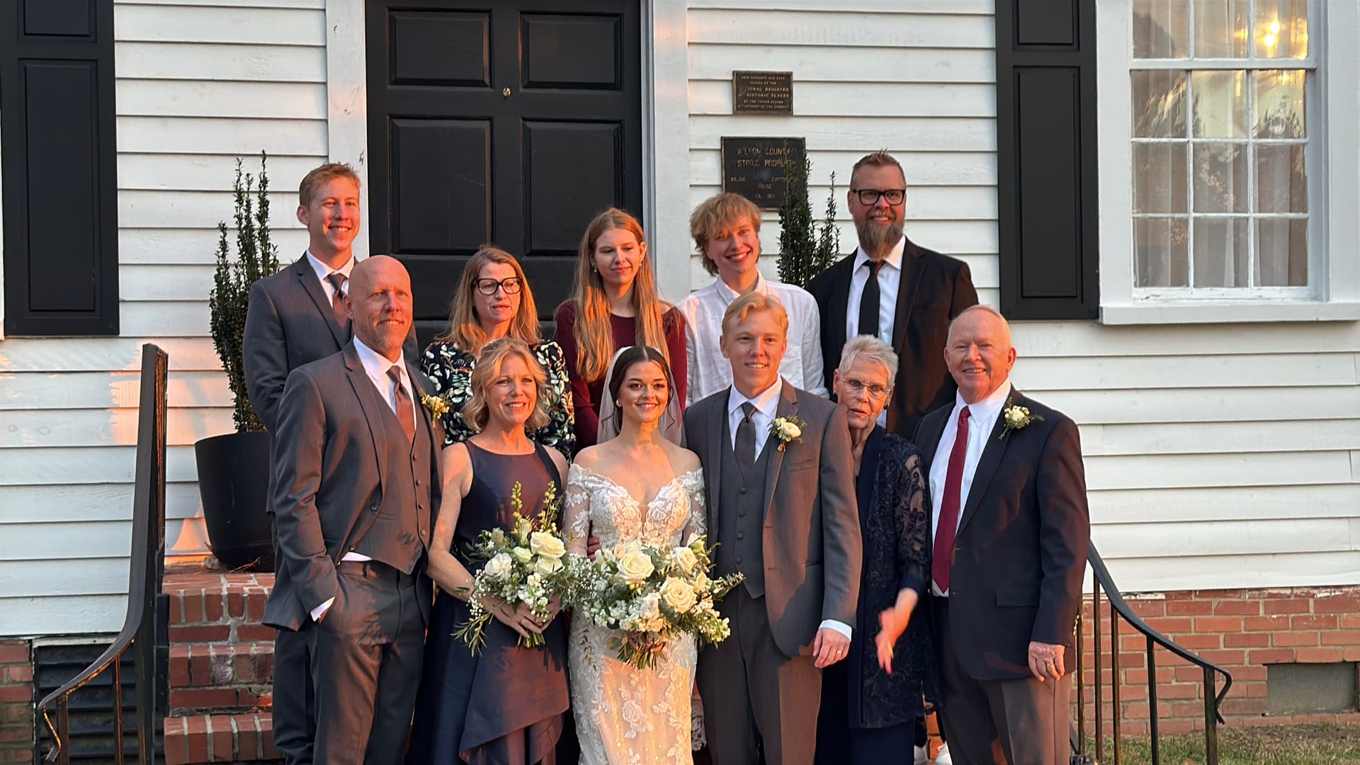 Bride and groom surrounded by smiling family members on the front steps of the 1821 Scarborough House historic manor at golden hour.