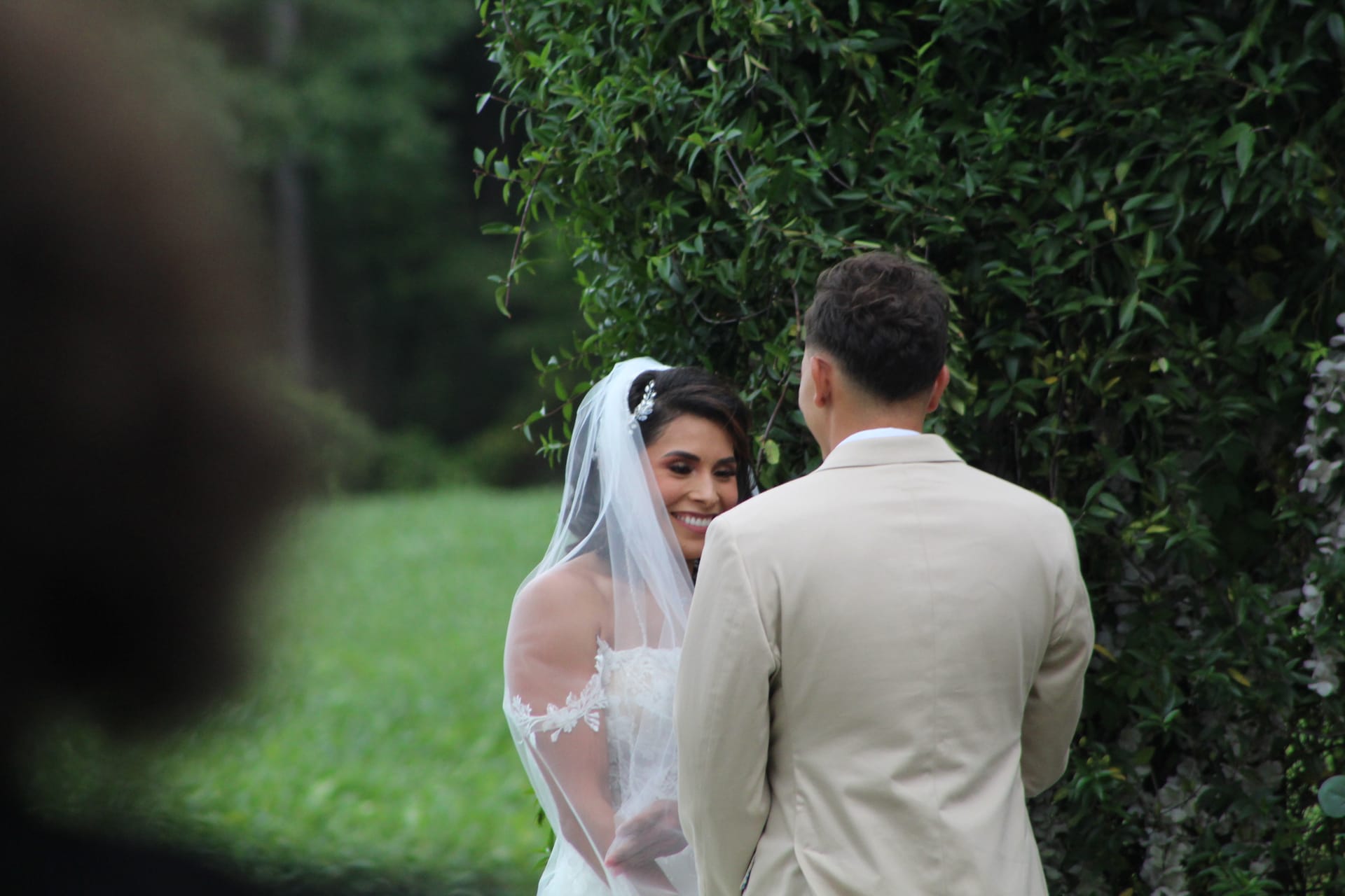 A smiling bride in a lace wedding gown and veil exchanges vows with her groom in a tan suit against a lush green garden hedge at Scarborough House.