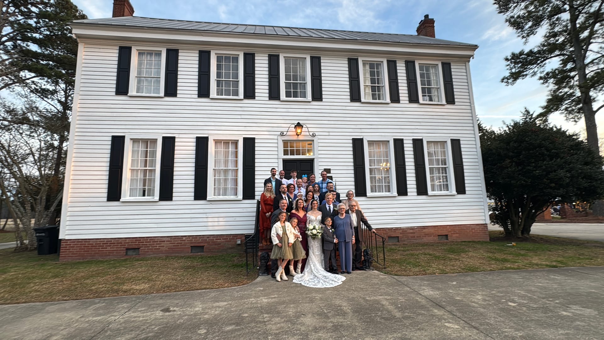 Wedding party and family gathered on the front steps of Scarborough House historic 1821 manor with white clapboard siding and black shutters at dusk.