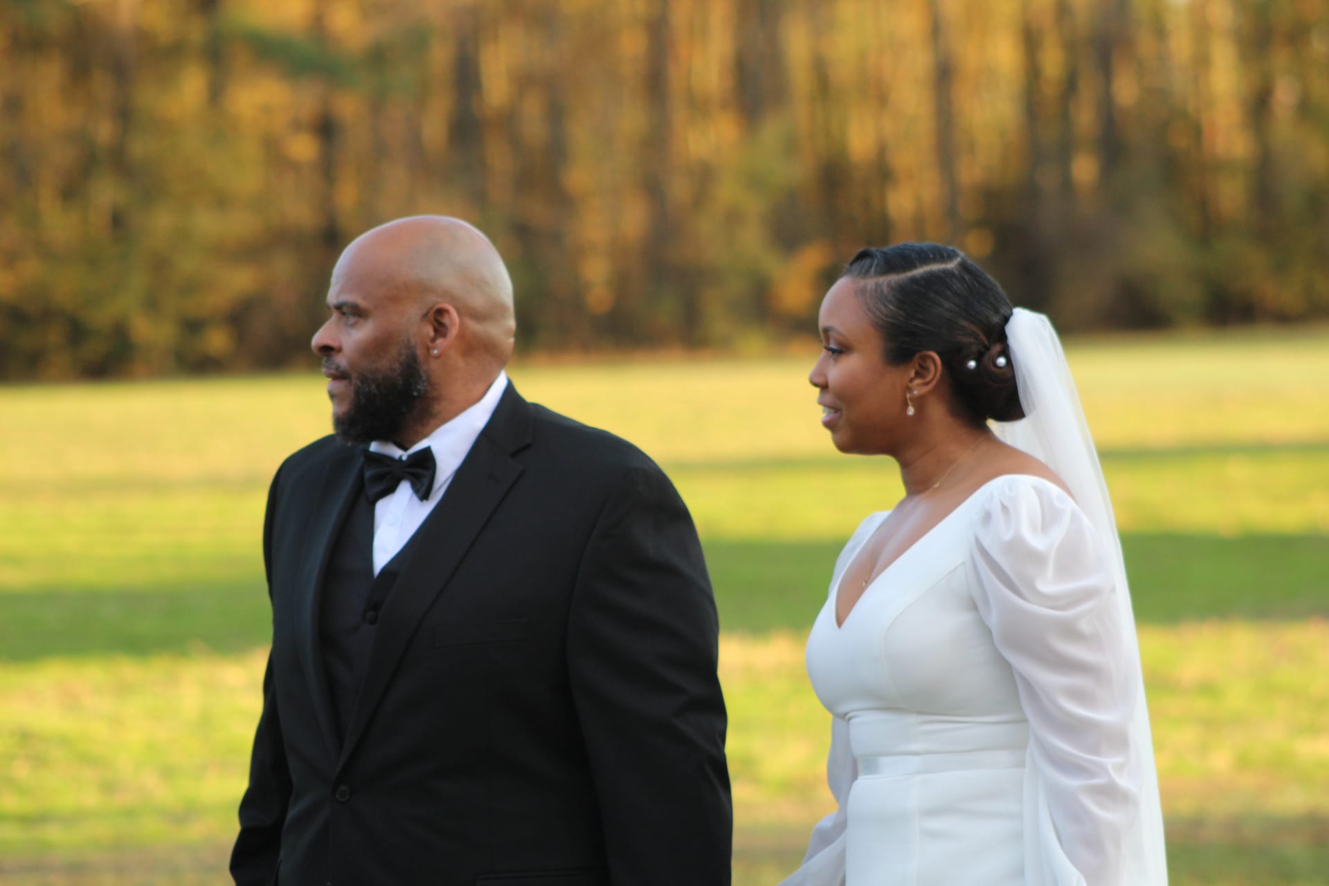 Bride in a white long-sleeve gown with veil and groom in a black tuxedo stand side by side outdoors against a warm autumn foliage backdrop at Scarborough House estate.