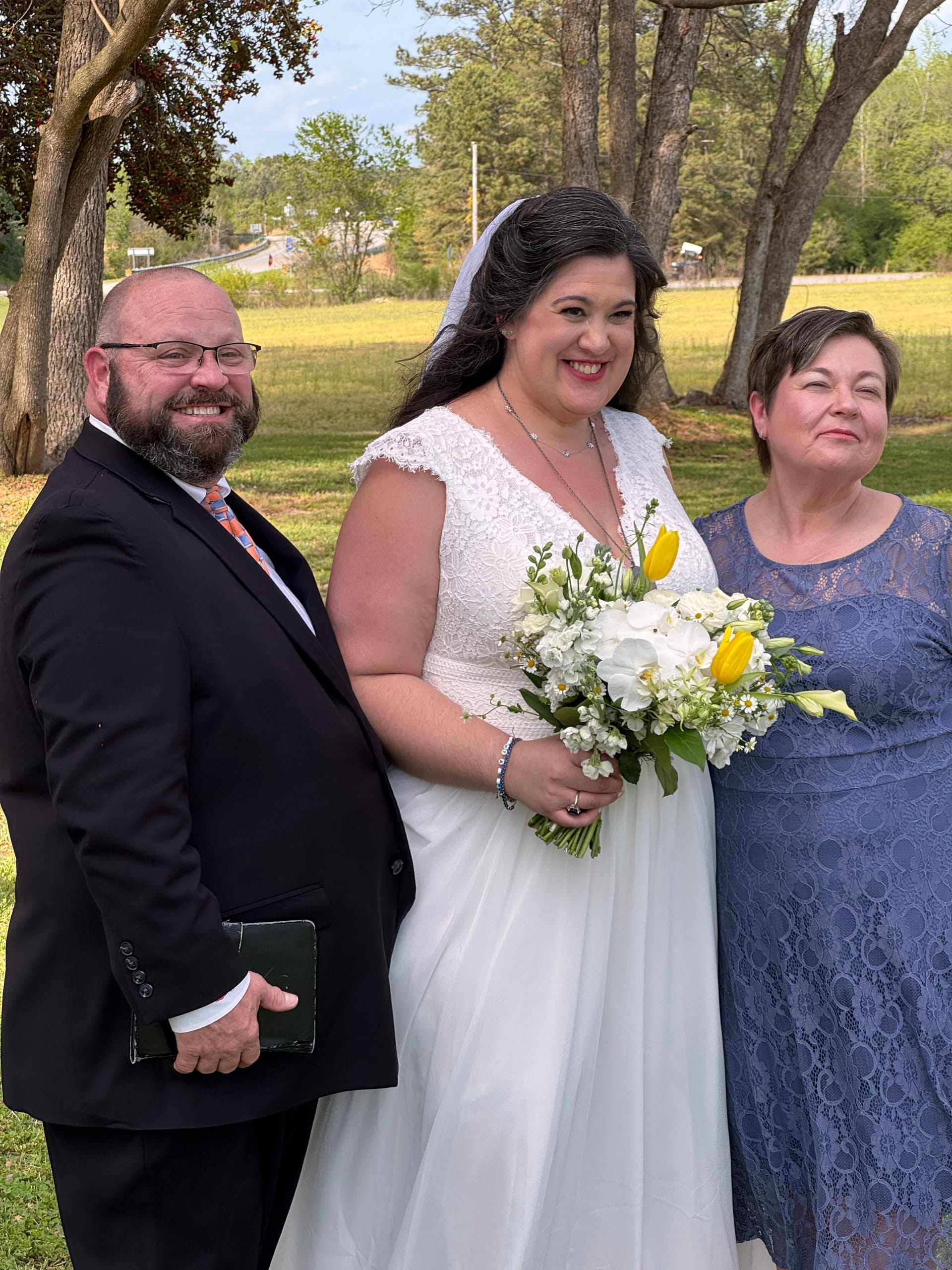 Smiling bride in a white lace gown holding a white and yellow bouquet stands between a bearded officiant in a black suit and a woman in a blue lace dress on the ceremony grounds at Scarborough House.