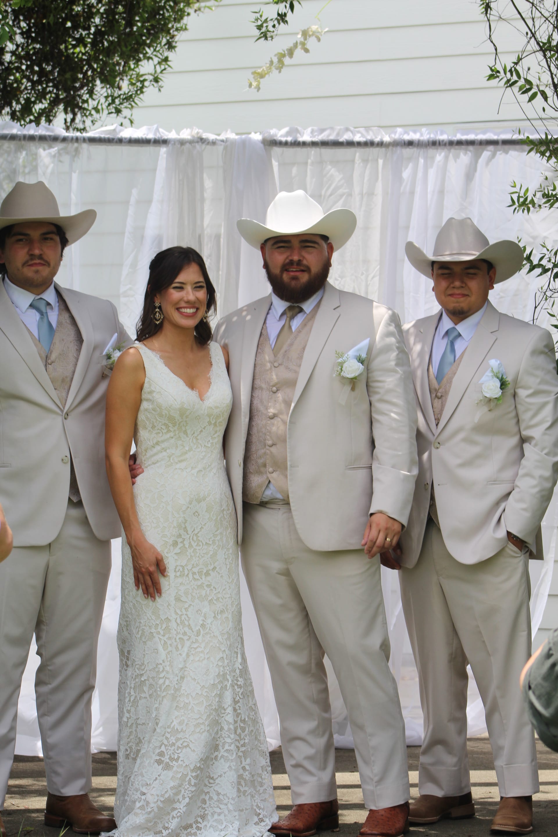 Bride in lace gown smiling alongside groom and two groomsmen in tan suits and cowboy hats against a white draped backdrop at an outdoor wedding ceremony.