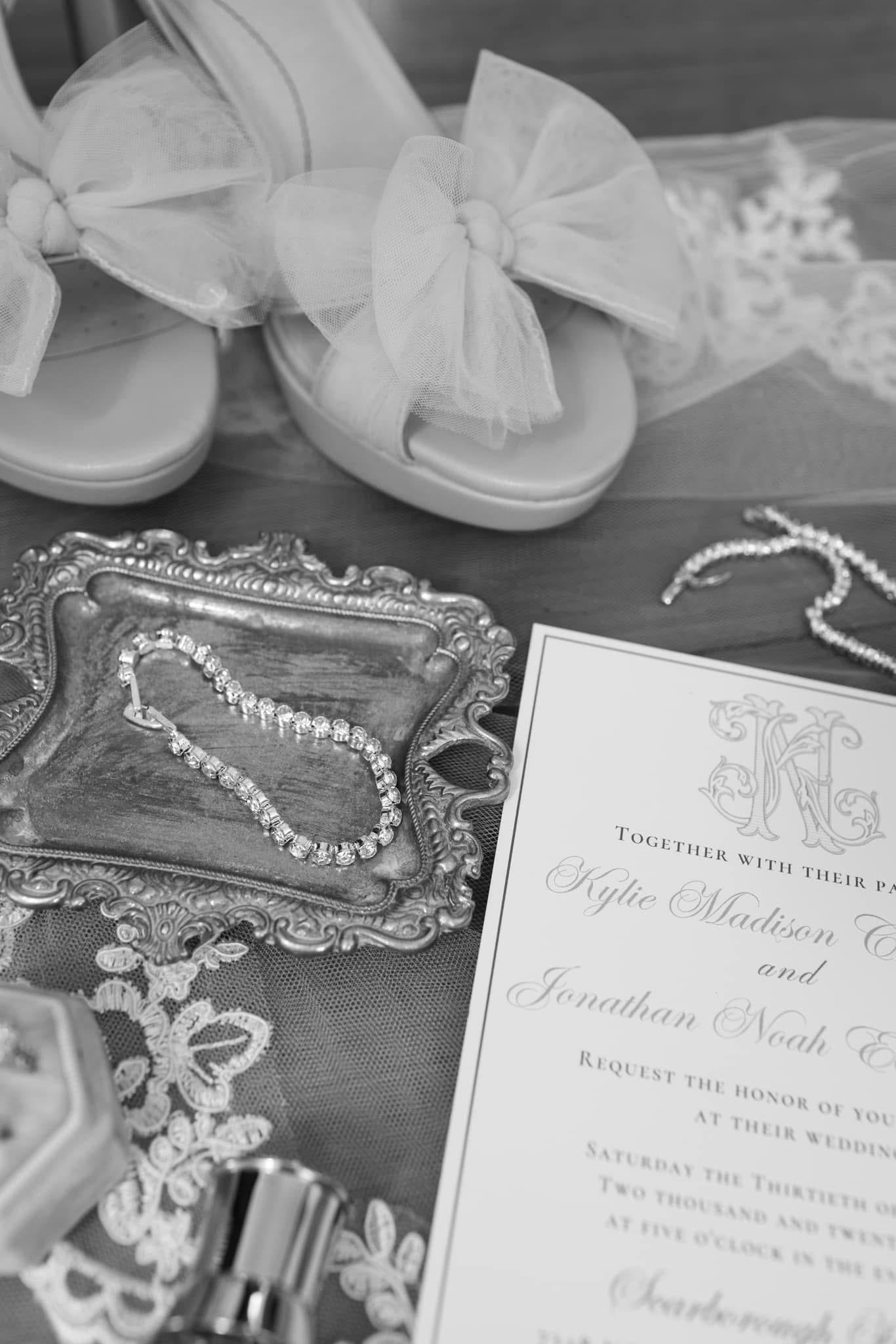 Black and white flat lay of bridal accessories at Scarborough House including a wedding invitation for Kylie Madison and Jonathan Noah, white heeled sandals with tulle bows, a diamond tennis bracelet on a silver ornate tray, and delicate lace veil.