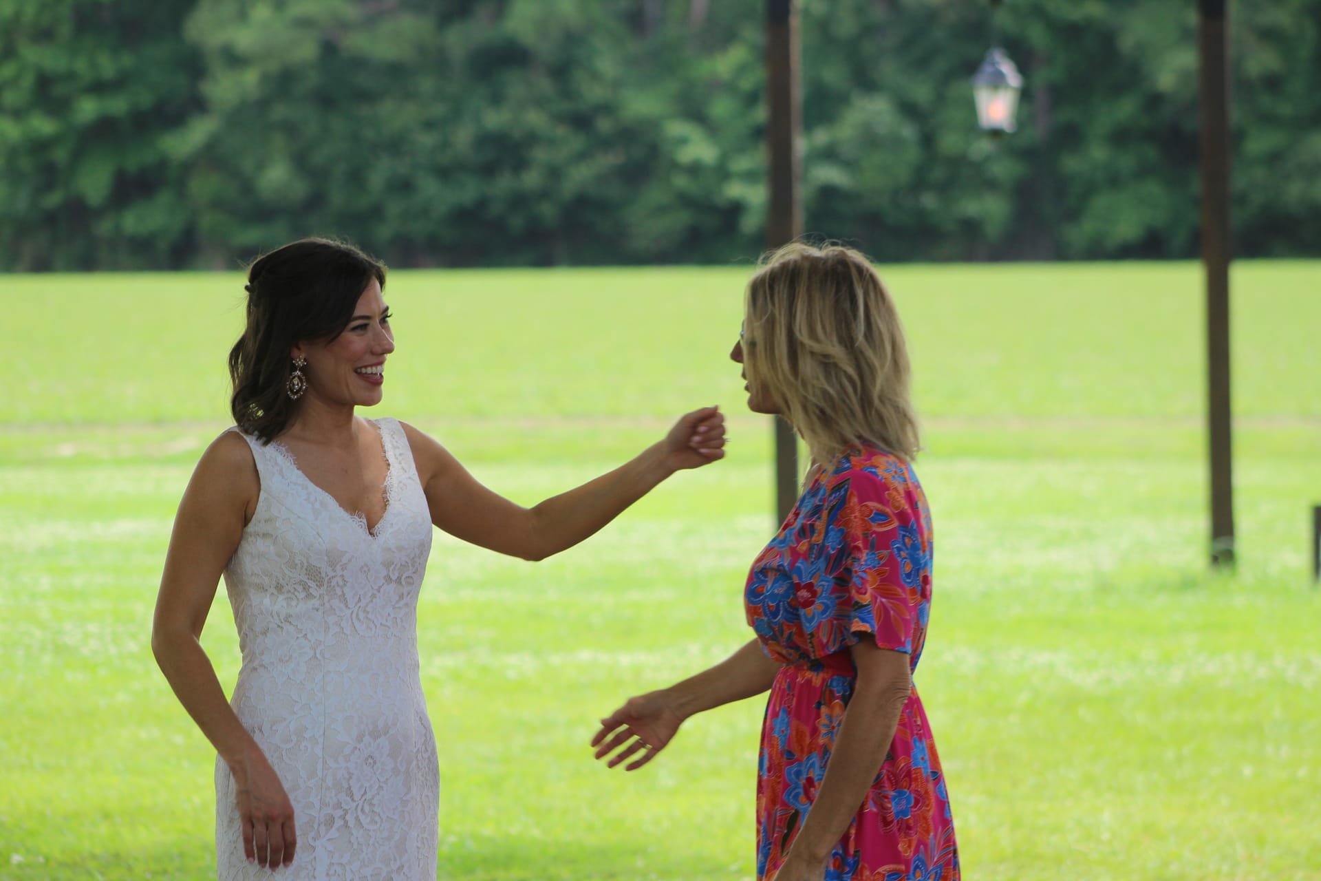Bride in a white lace gown laughs and reaches toward a blonde guest in a colorful floral dress on the sunny green lawn at Scarborough House wedding venue.