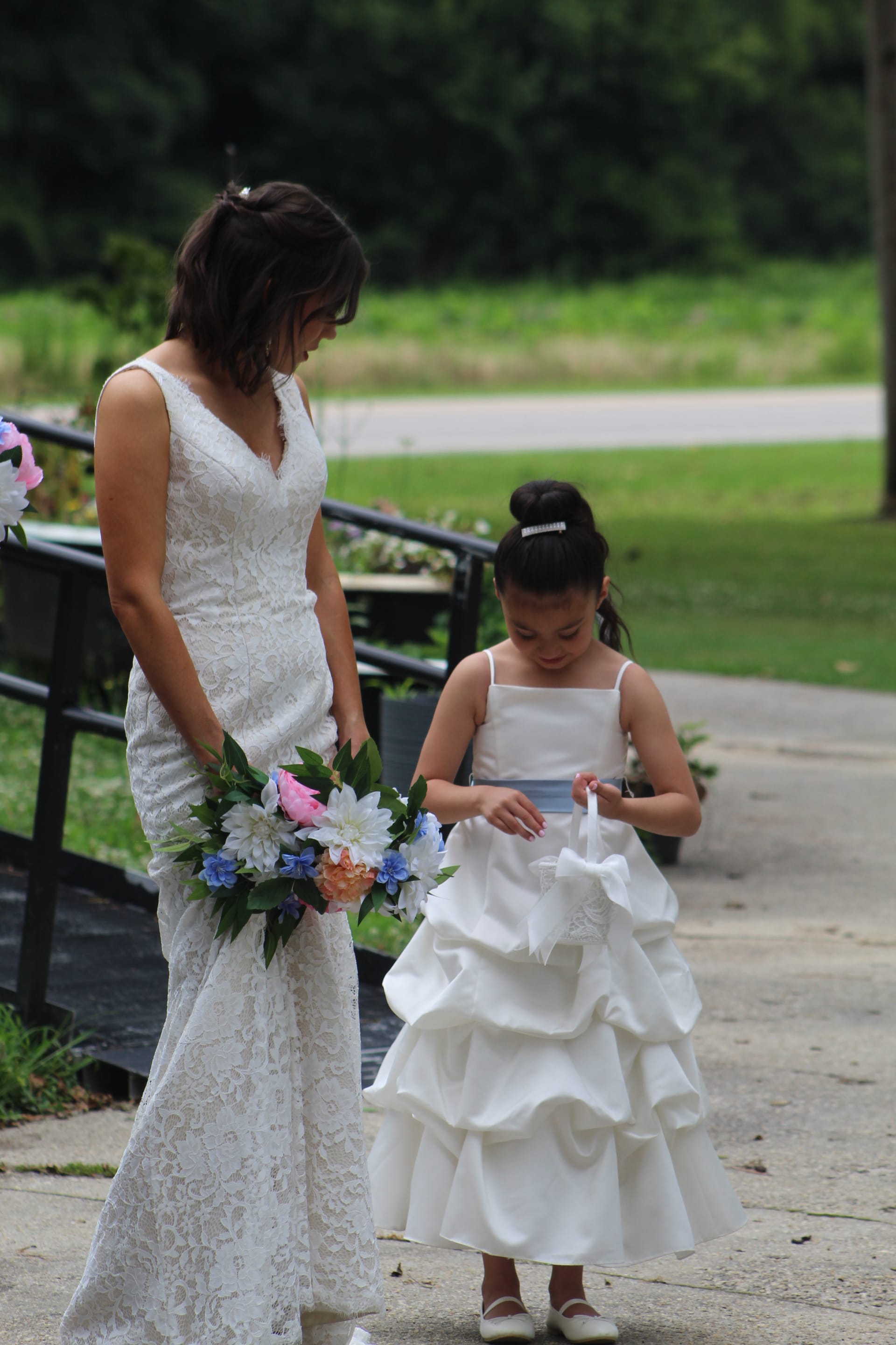 Bride in lace wedding gown holding a colorful bouquet looks down at a young flower girl in a white ruffled dress on a paved garden path surrounded by greenery.