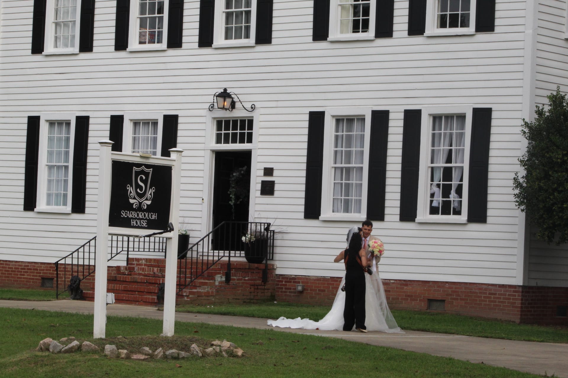 Bride and groom share a romantic dip kiss on the front lawn of Scarborough House historic wedding venue with the white 1821 manor and marquee sign visible behind them.