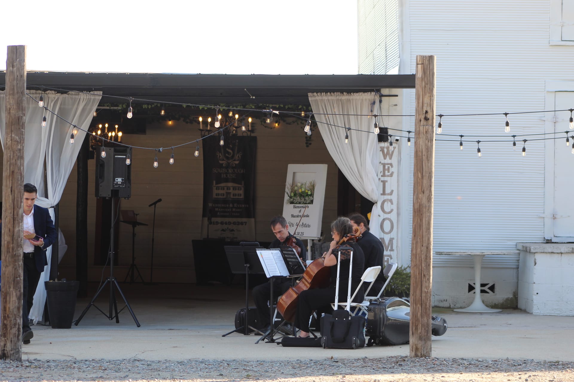 A string ensemble performs beneath warm Edison bulb lights and flowing white drapes at the entrance of Scarborough House wedding venue at dusk.