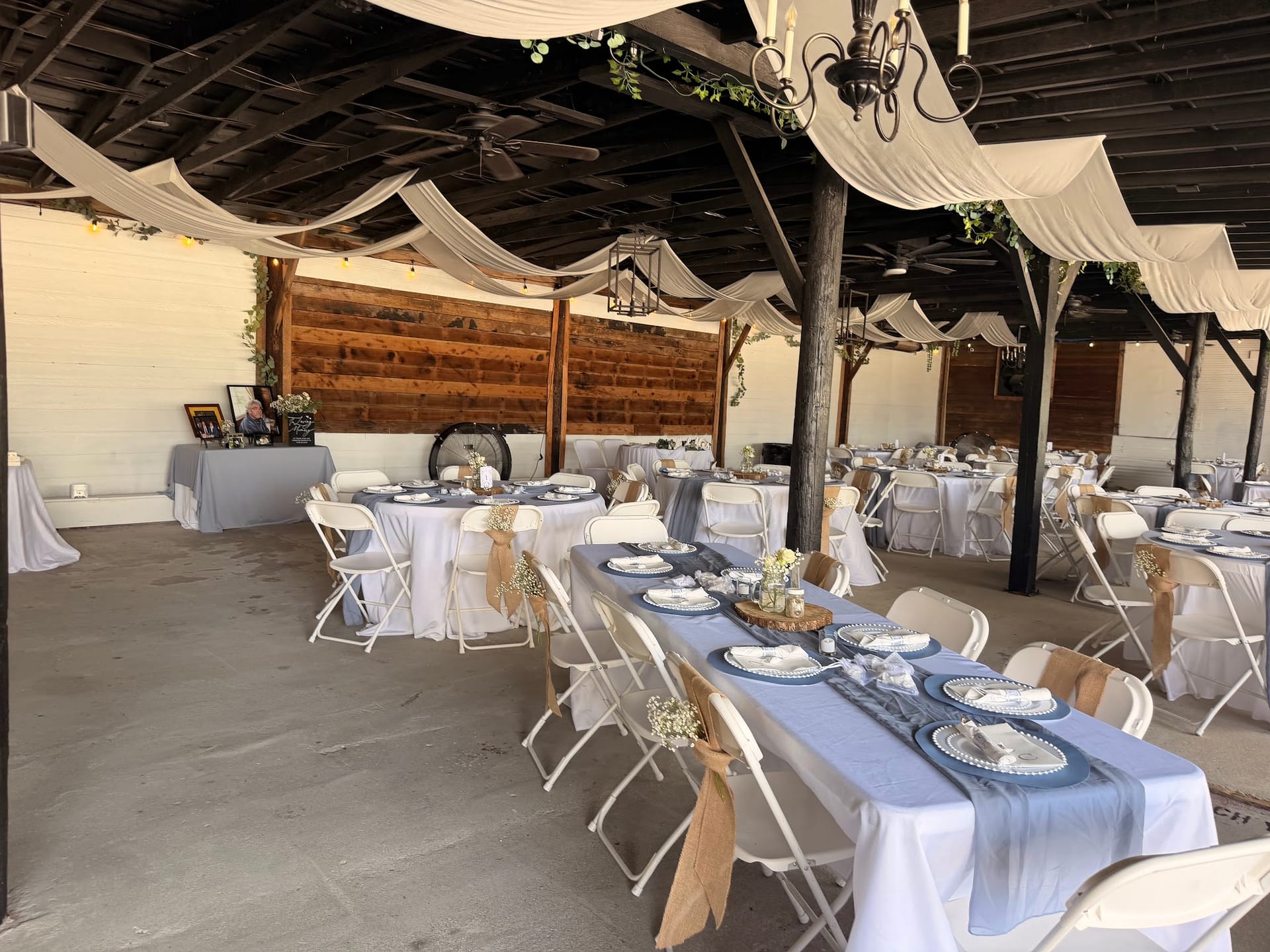 Elegantly styled outdoor reception pavilion at Scarborough House featuring white draped ceiling, chandelier, white folding chairs, and tables set with dusty blue runners and floral centerpieces against a rustic wood-panel backdrop.