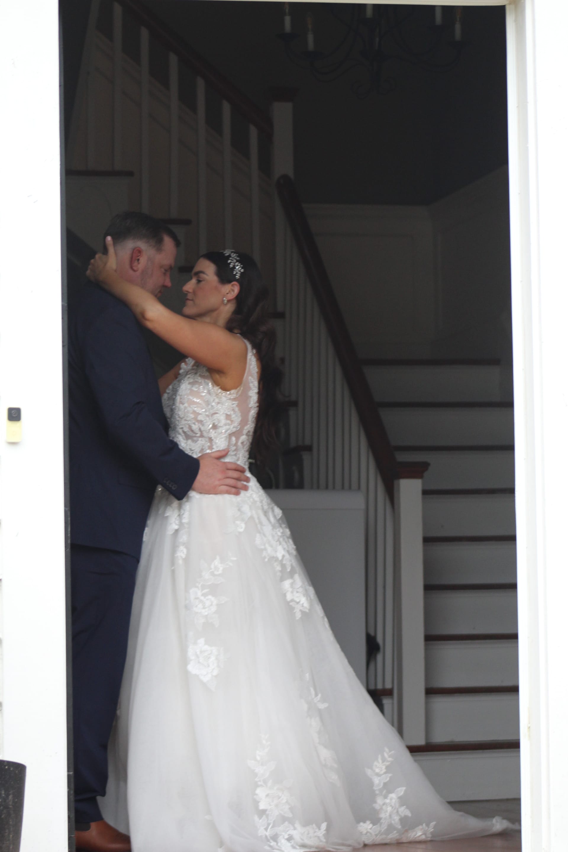 Bride in a floral lace ballgown and groom in a navy suit share an intimate embrace in the grand hall doorway framed by a sweeping staircase at Scarborough House.