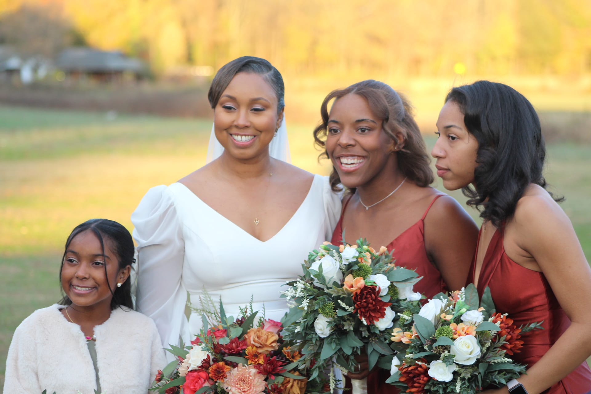 Bride in a white gown smiles with two bridesmaids in rust-red dresses and a young girl in cream, all holding lush autumn bouquets of white roses, dahlias, and greenery against a golden-hour farmland backdrop at Scarborough House.