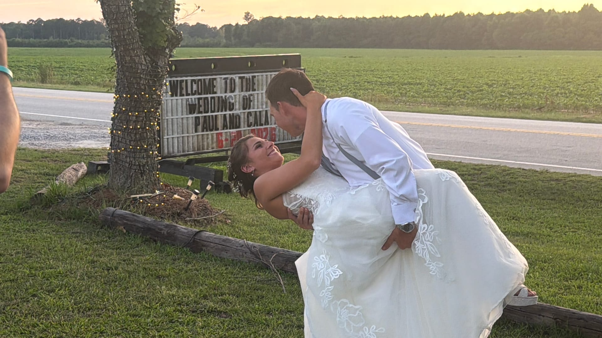 Groom dips and kisses his bride in front of the Welcome to the Wedding of Paul and Cala marquee sign at golden hour with open farmland fields in the background.
