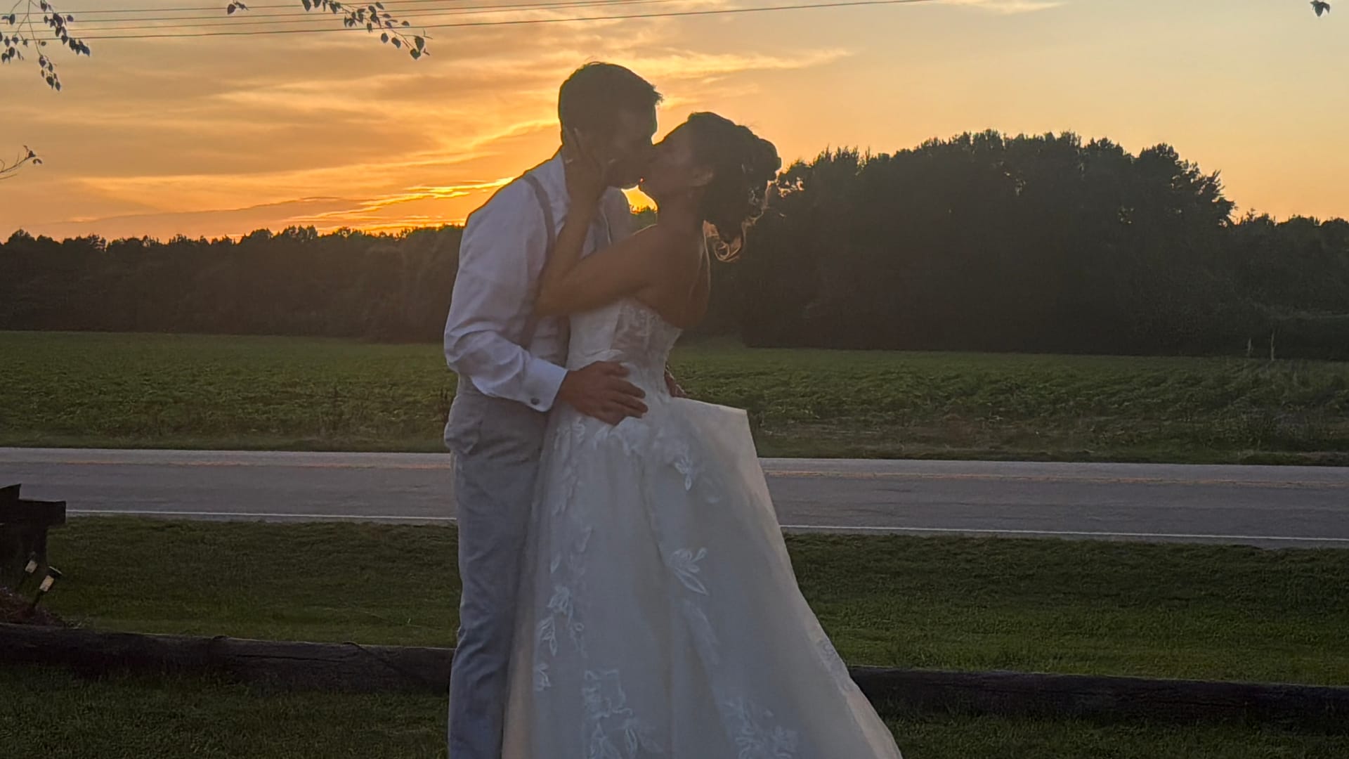 Bride and groom share a romantic kiss silhouetted against a glowing golden sunset with rolling farmland fields and tree line behind Scarborough House estate.