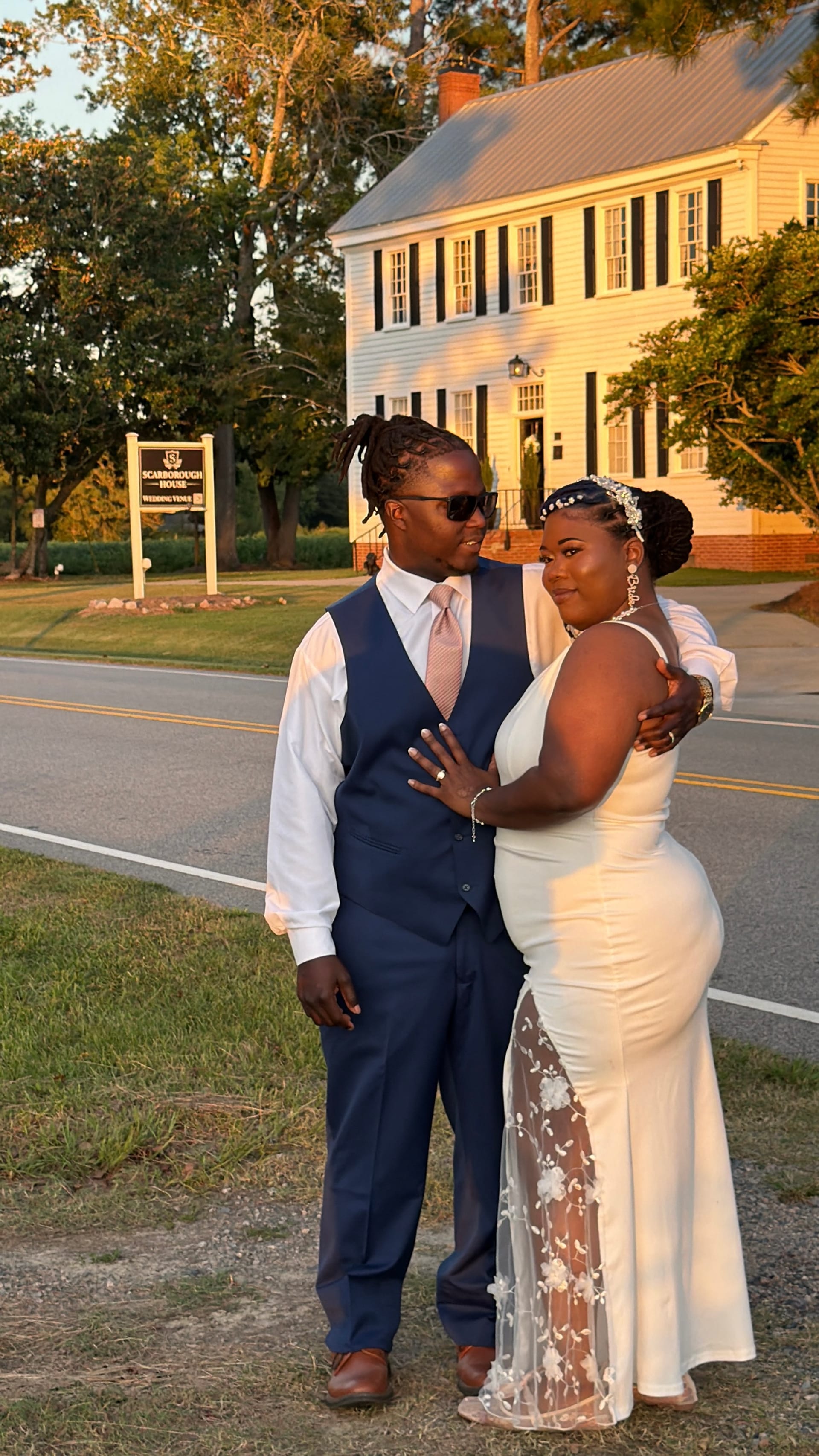 Bride in a white fitted gown and groom in a navy suit embrace in front of the Scarborough House wedding venue sign at golden hour with the 1821 historic manor glowing in the background.