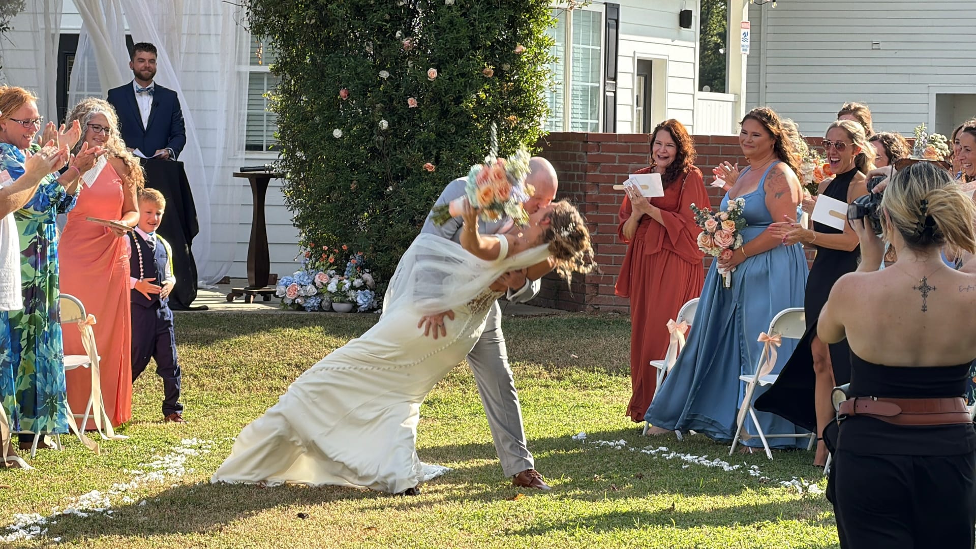 Groom dips bride for a dramatic kiss at the end of their outdoor wedding ceremony on the sun-drenched ceremony lawn at Scarborough House as guests cheer and applaud around them.