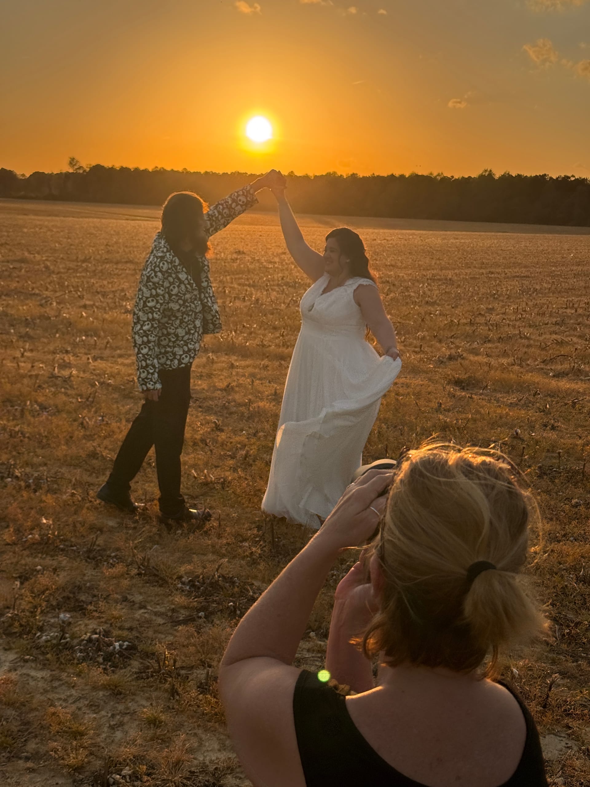 A couple dances together in an open farmland field at golden hour as a photographer captures the moment against a brilliant orange sunset sky.