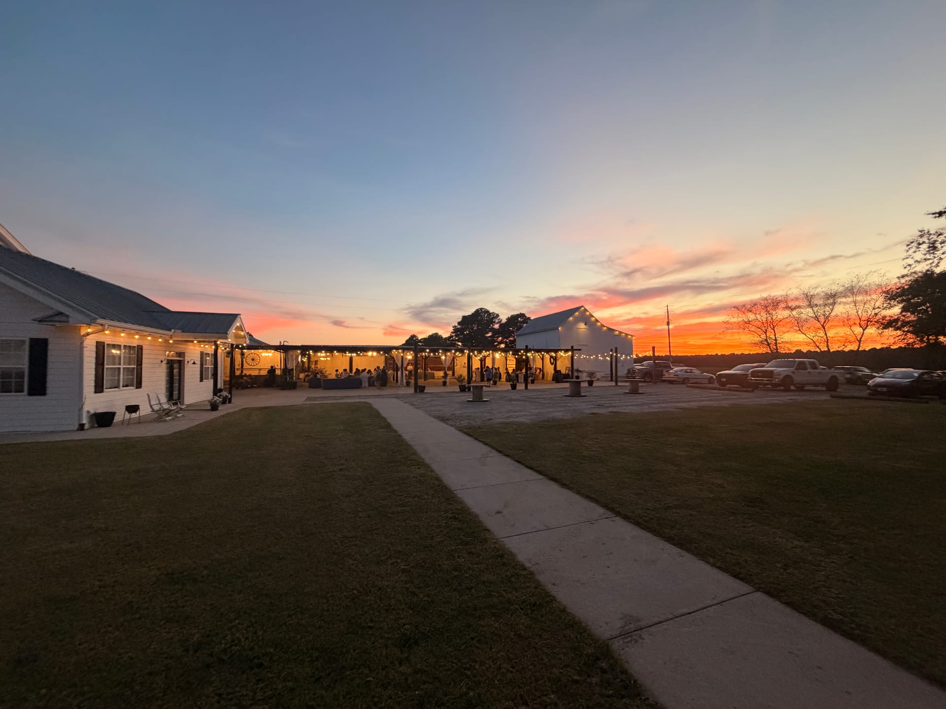 Warm sunset glows orange and pink over Scarborough House historic estate venue with string-lit covered pavilion and white grand hall at dusk during an outdoor reception.