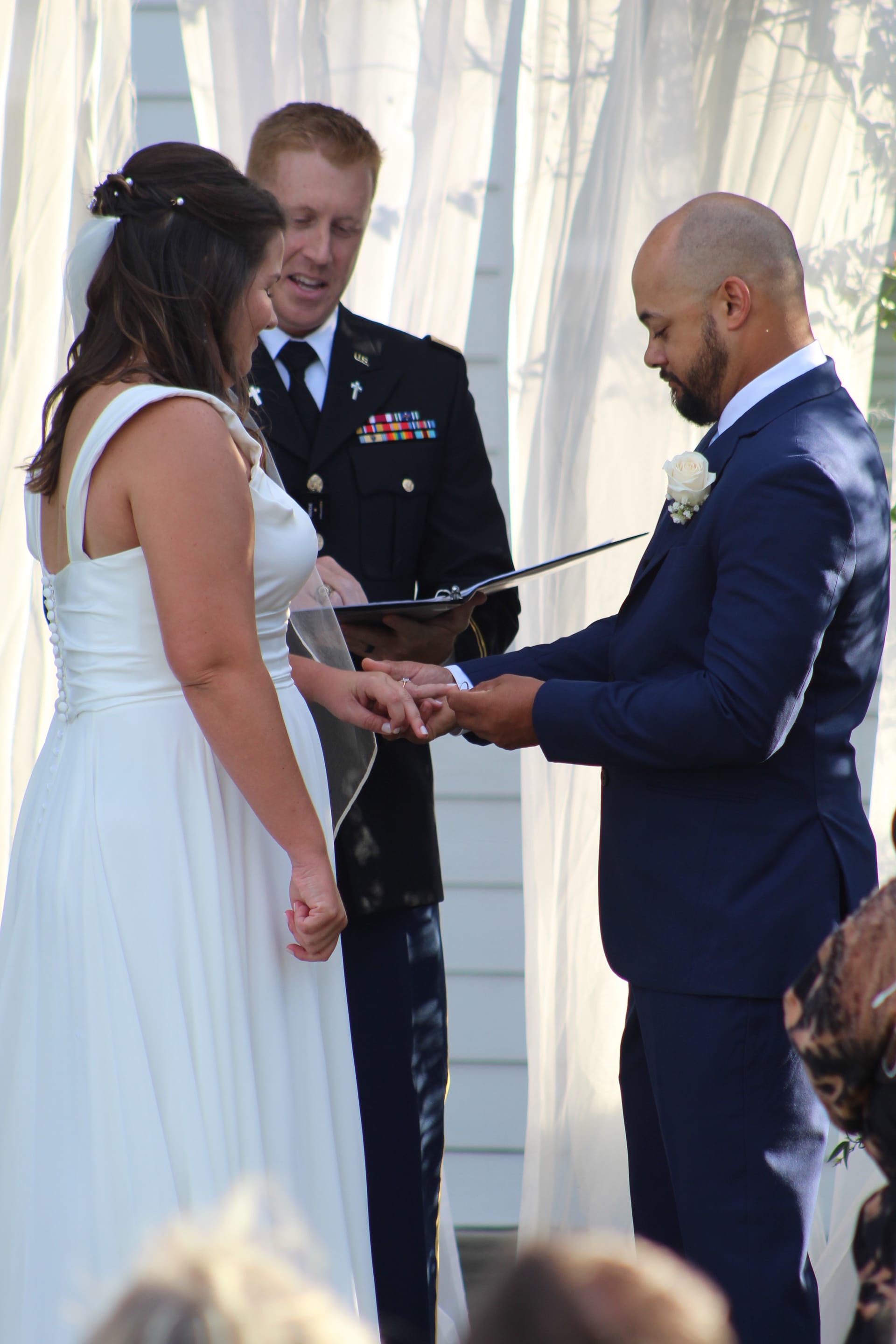 Groom in navy suit places wedding ring on brides finger during outdoor ceremony officiated by a military chaplain beneath white draped curtains at Scarborough House.