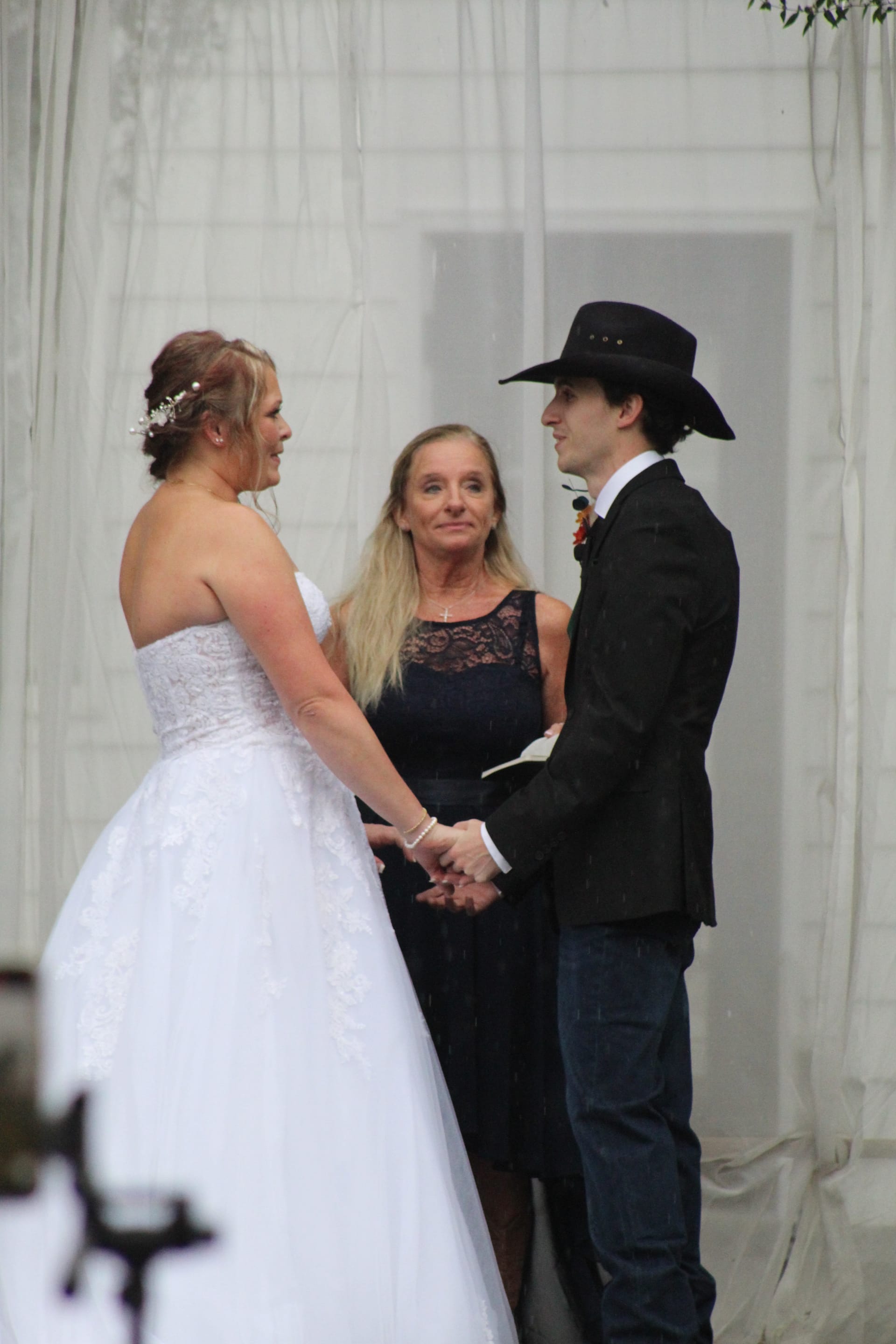 Bride in a white strapless gown and groom in a black cowboy hat and blazer hold hands during their wedding ceremony officiated by a woman in a navy lace dress at Scarborough House.