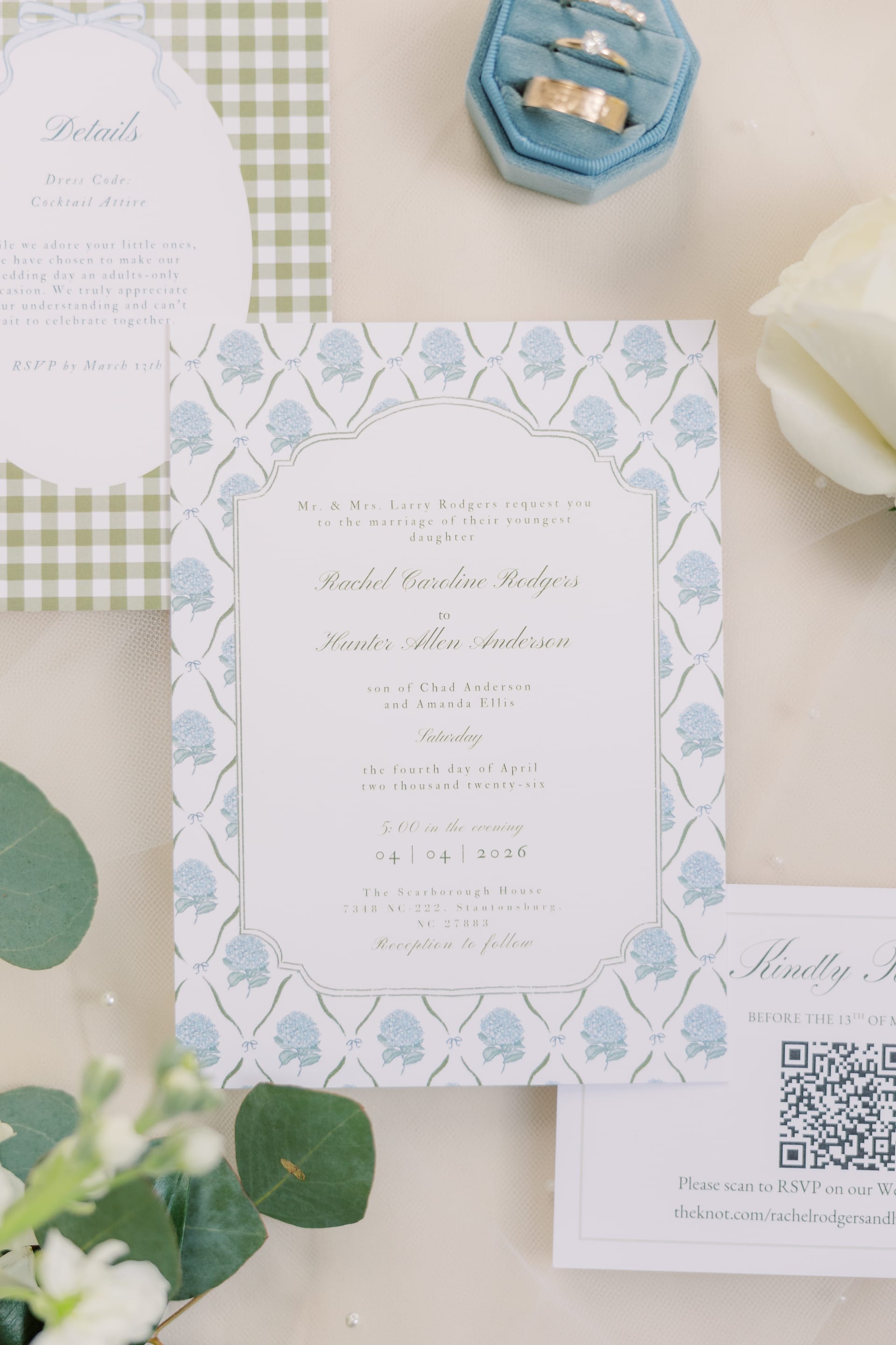 Elegant wedding invitation suite with blue hydrangea border, gold rings in a blue velvet box, and white flowers.