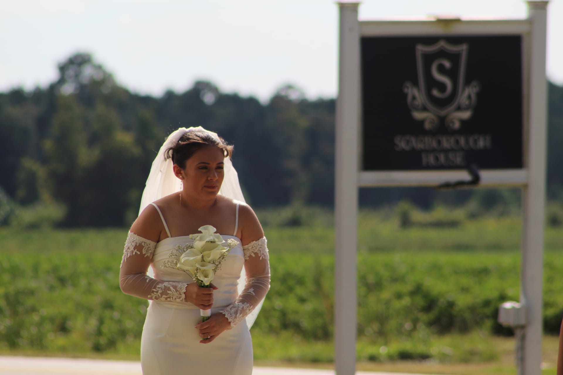 Bride in a white off-shoulder lace gown holding a white calla lily bouquet standing near the Scarborough House estate sign with green fields and trees in the background.